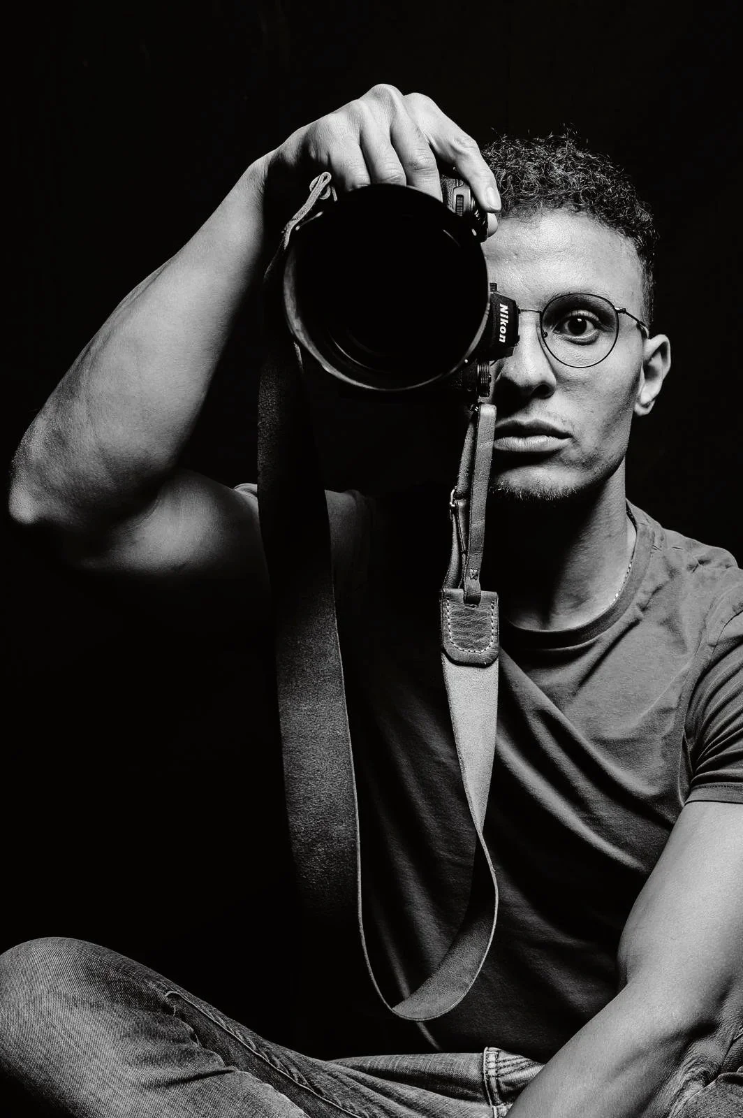 Ritratto di un uomo con occhiali che si fa un autoritratto con una fotocamera Nikon, in bianco e nero, su sfondo nero.