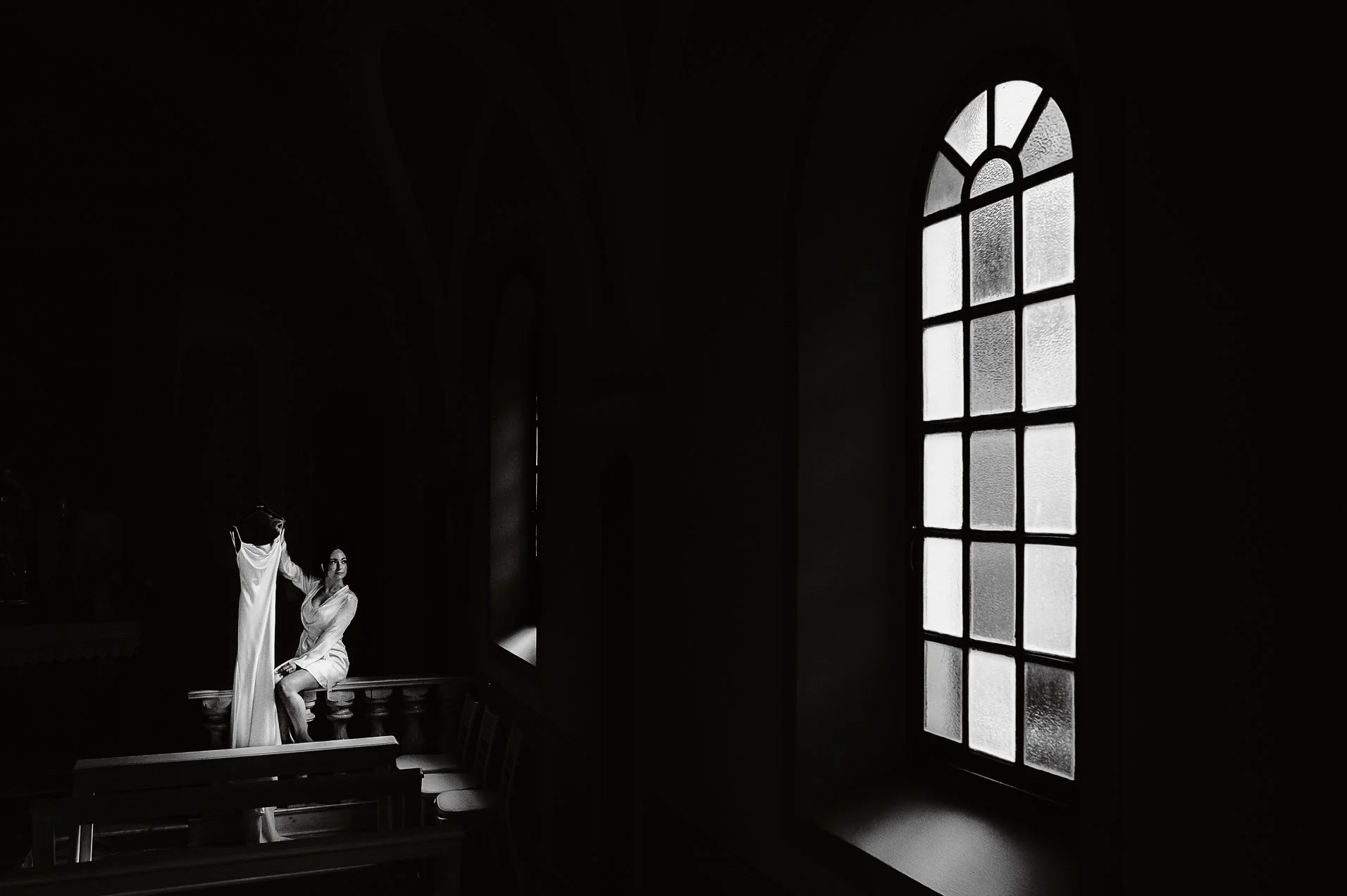 Interno di chiesa con grande finestra di vetro opaco e donna seduta su una panca, che guarda fuori, vestita con abito chiaro. La scena è in bianco e nero.