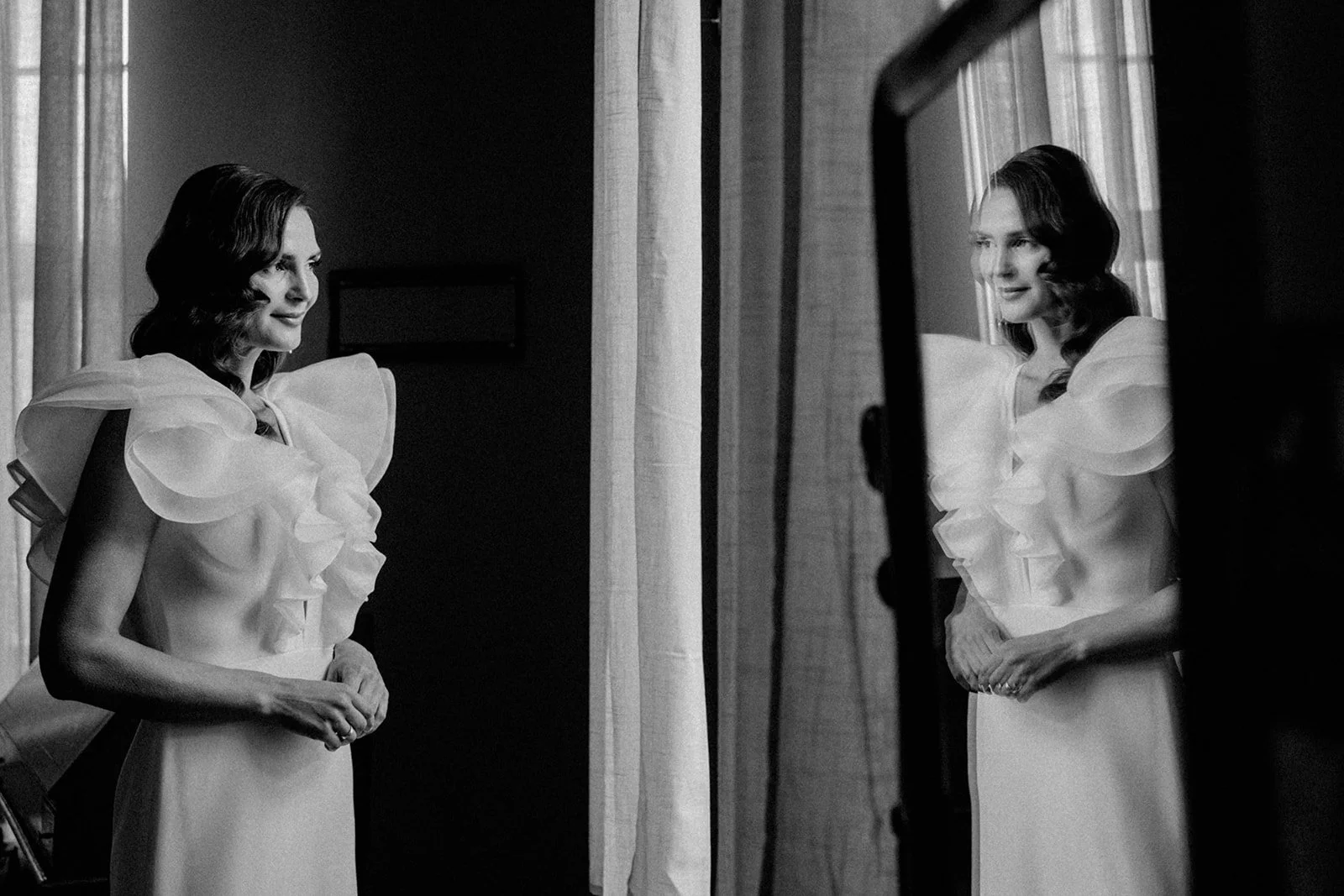 Schwarz-weiß Fotografie einer Frau, die sich im Spiegel betrachtet, mit eleganter Frisur und einem Kleid mit großen, gepufften Schultern, vor einer Fensterbank.