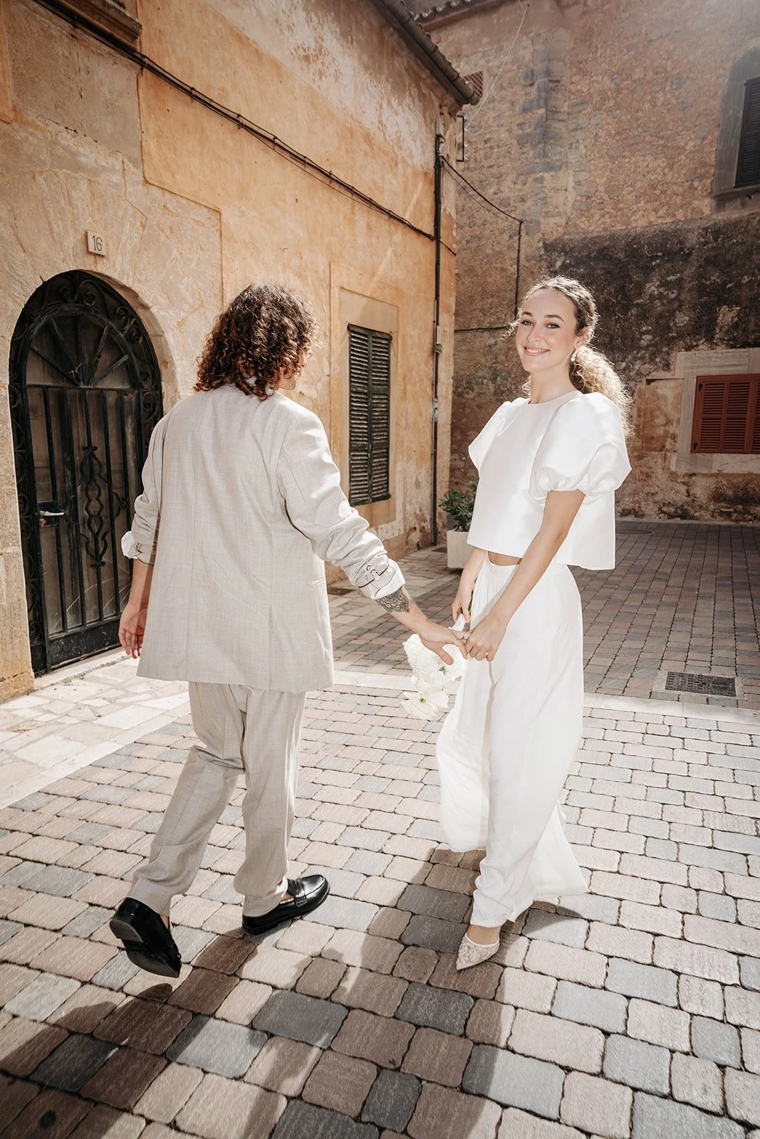Zwei Frauen in weißen Kleidern halten sich an den Händen, draußen auf einem Kopfsteinpflaster, eine lächelnd, die andere von hinten sichtbar.