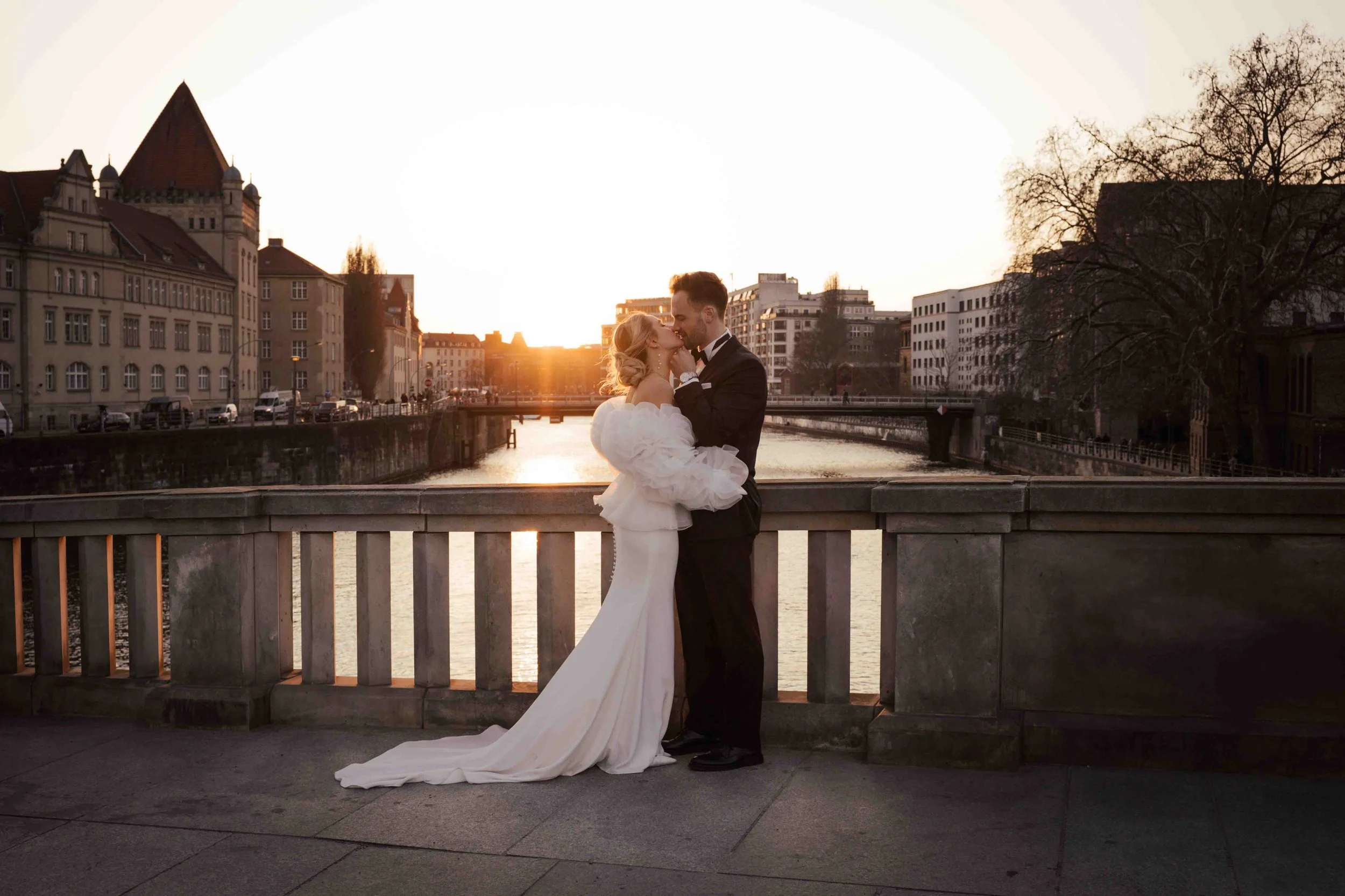 Ein elegantes Brautpaar beim Fotografieren auf einer Brücke bei Sonnenuntergang in einer urbanen Umgebung mit Wasser, alten Gebäuden und einem Baum im Hintergrund.