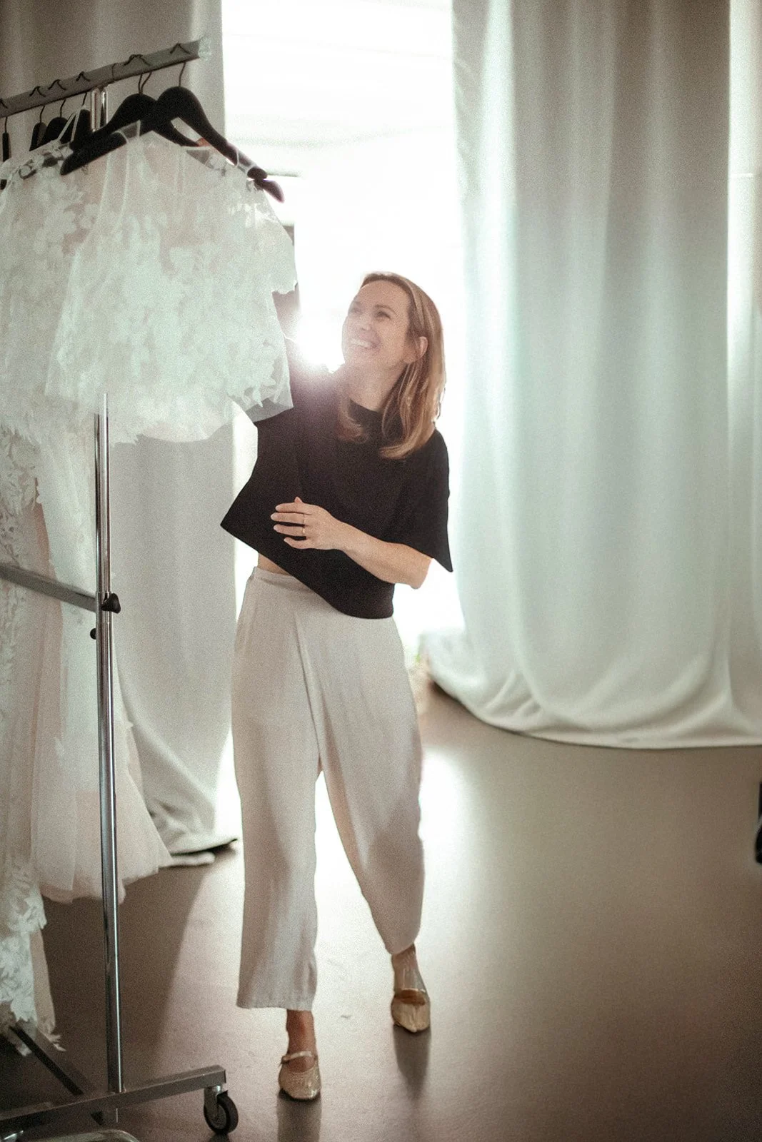 Eine lachende Frau sortiert weiße Brautkleider auf einer Kleiderstange in einem hell erleuchteten Studio.