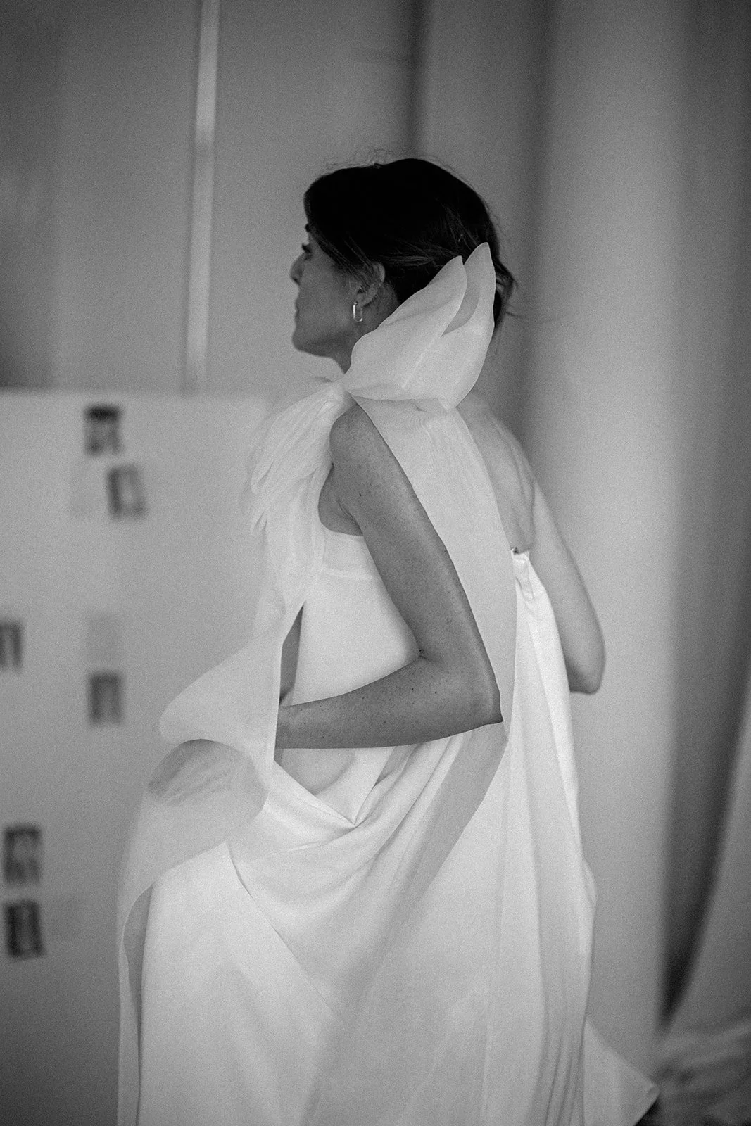 Schwarz-Weiß-Foto einer Frau in einem weißen Kleid mit großer Schleife, die nach hinten gewandt ist und seitlich steht.
