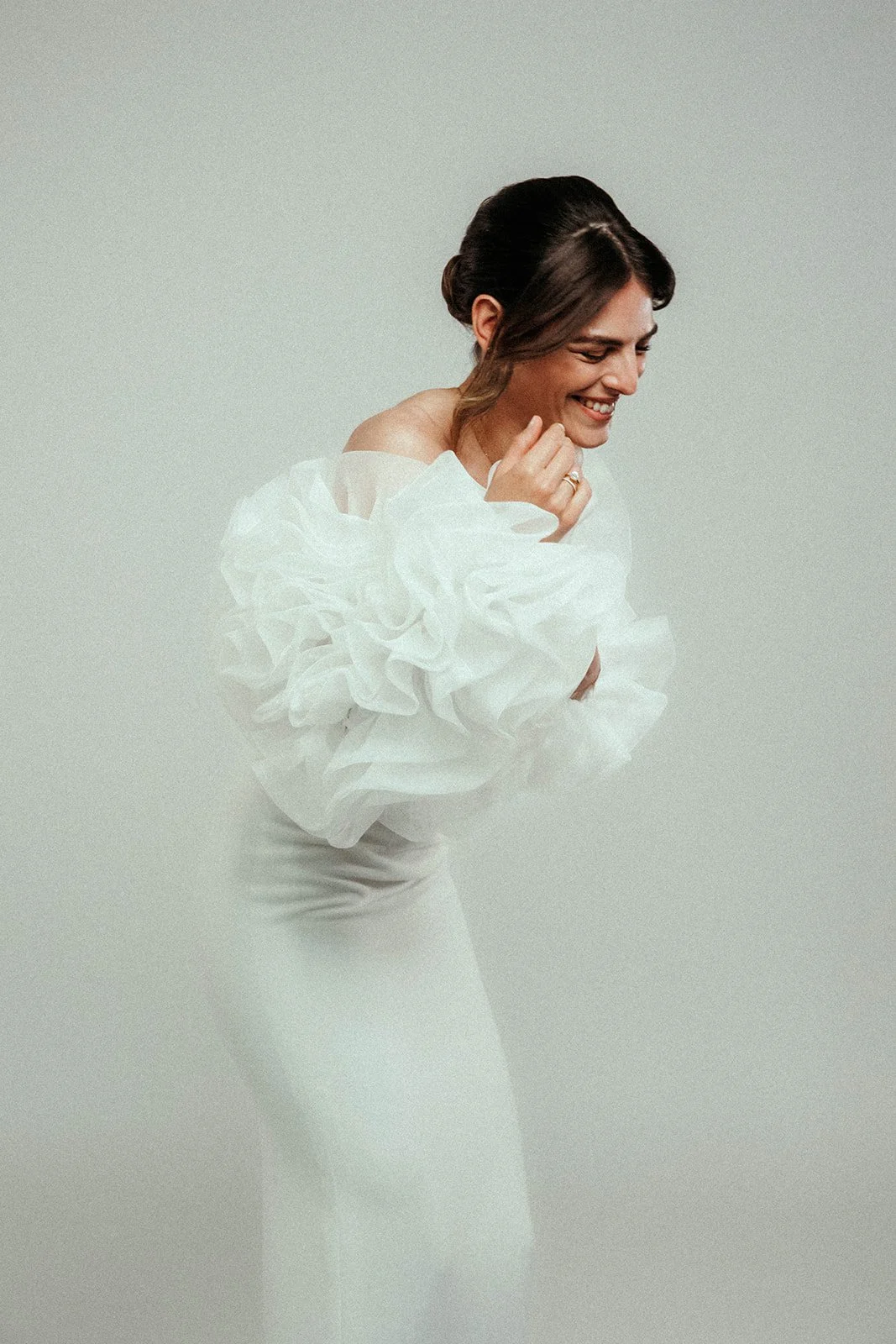 Model trägt ein weißes Hochzeitskleid mit großen Rüschen, lächelt und scheint fröhlich zu sein, vor einem hellen, einfarbigen Hintergrund.