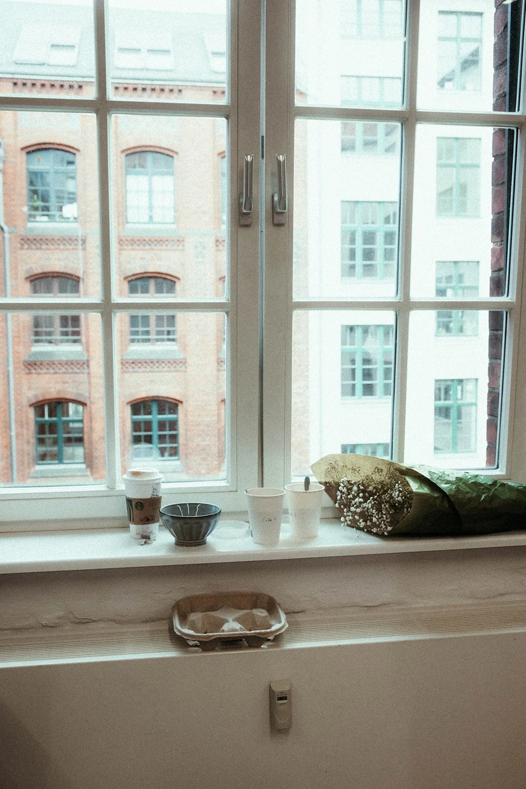 Fensterbank mit Kaffeebecher, Blumenstrauß, leeren Schüsseln und Pappschachtel vor einem großen Fenster, dahinter Hochhäuser im Stadtbild.