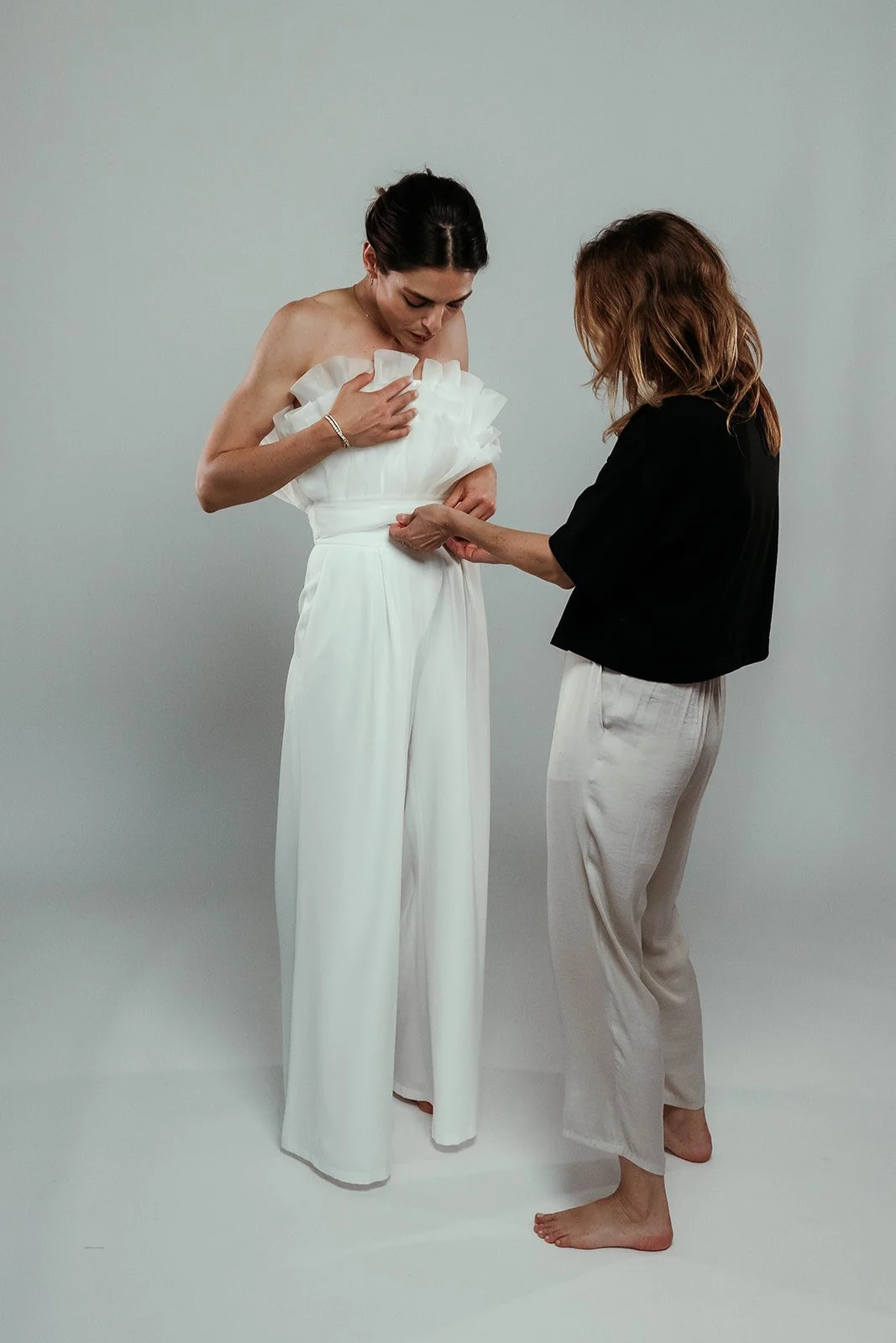 Einer Frau in einem eleganten weißen Kleid wird von einer Stylistin in schwarzen Oberteil und weißen Hosen geholfen, das Kleid anzuprobieren.