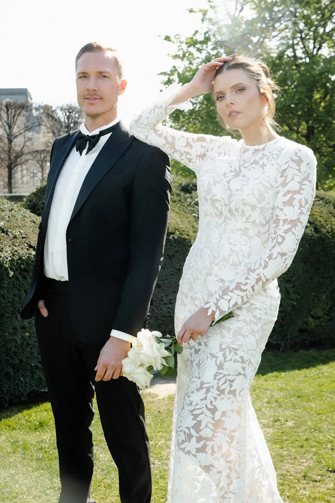 Ein Paar in formeller Kleidung, ein Mann in einem schwarzen Anzug mit Fliege und eine Frau in einem weißen, hochgeschlitzten Brautkleid mit Blumendesign, im Freien bei sonnigem Wetter.