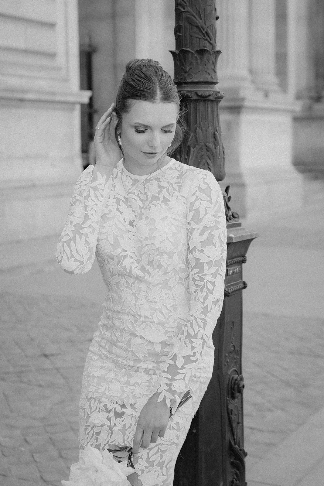 Schwarz-weiß-Foto einer Frau in einem langen, blumenbestickten Kleid, die an einem Laternenpfahl lehnt und ihre Hand an den Kopf hält.