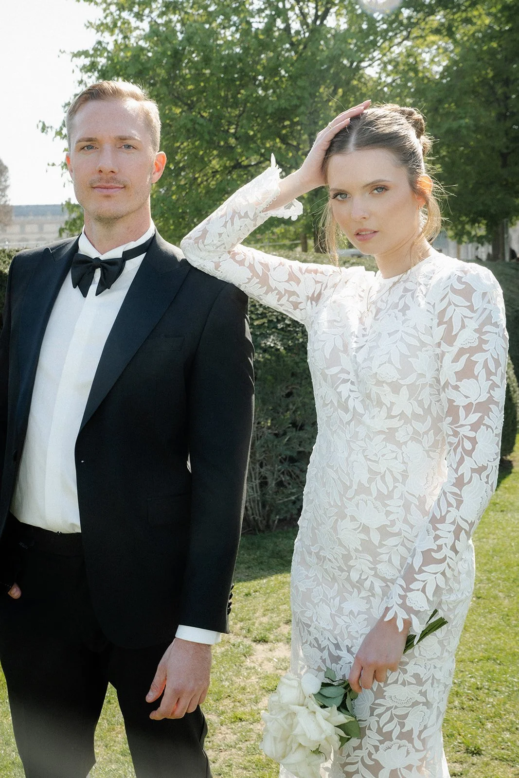 Ein Mann in einem schwarzen Anzug mit Fliege und eine Frau in einem weißen, floralen Brautkleid stehen im Freien mit grünen Bäumen und Gras im Hintergrund.