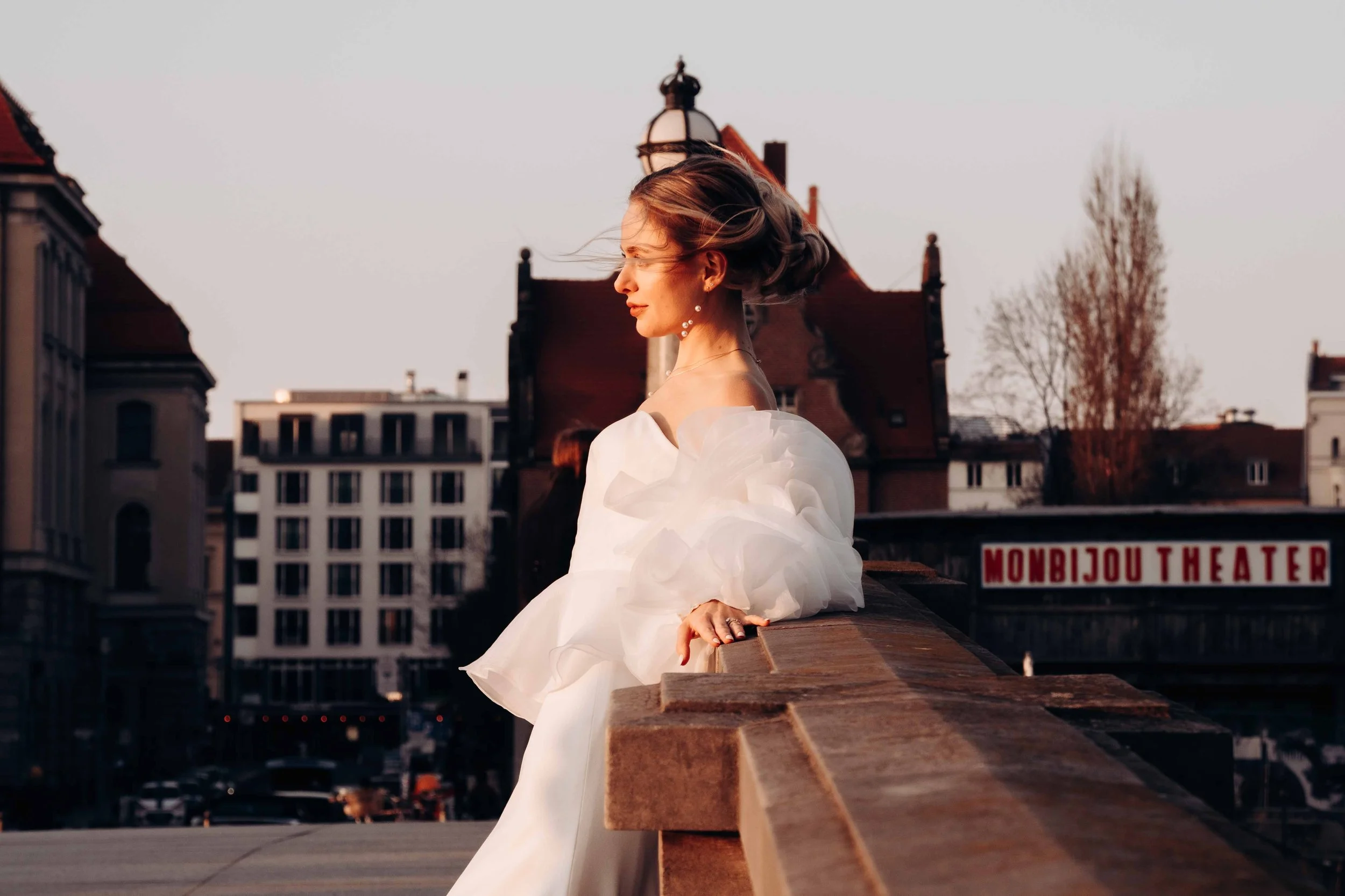 Frau in einem weißen Kleid mit voluminösen Stoffärmeln, die auf einer Brücke in der Stadt sitzt, bei Sonnenuntergang.