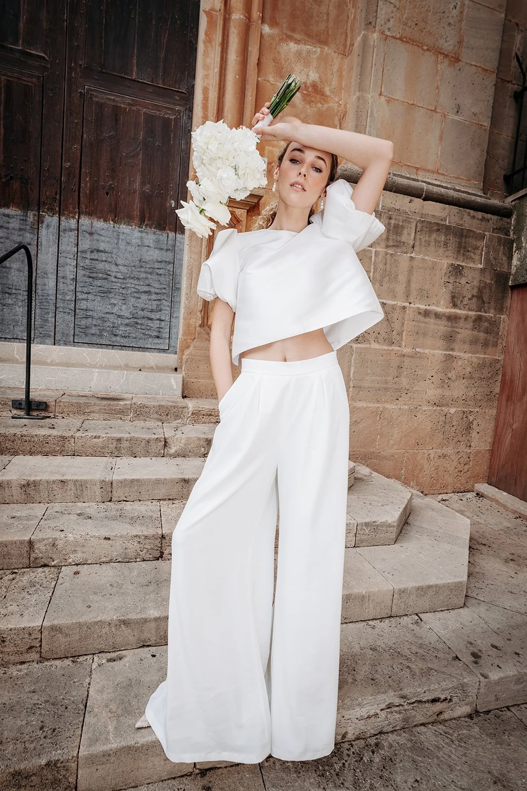 Junge Frau in weißen Turnwear-Mode mit lockigem Haar, die auf den Stufen einer historischen Steinarchitektur steht und eine weiße Blumensträuße hält.