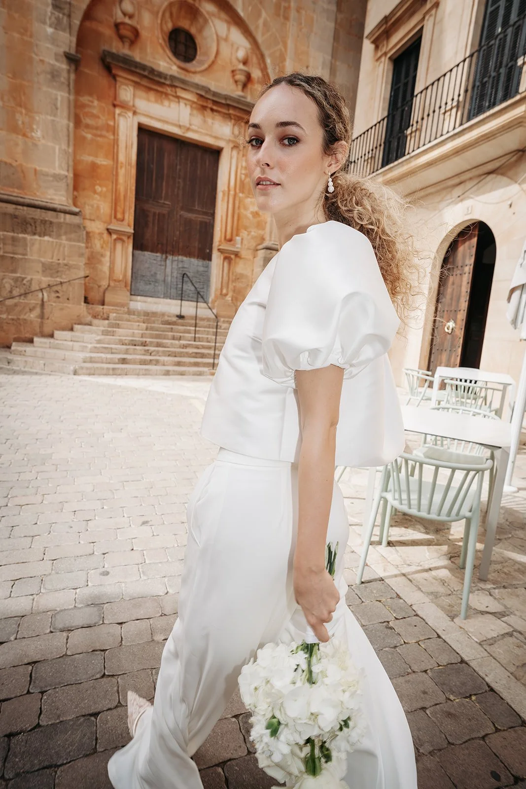 Junge Frau in weißem Outfit hält einen großen Blumenstrauß in der Hand im Freien auf gepflastertem Boden, mit alten Gebäuden im Hintergrund.