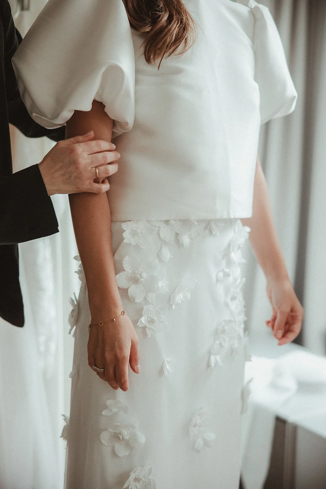 Nahaufnahme einer Person in einem weißen Brautkleid mit Blumenverzierungen, jemand anderes richtet das Kleid.
