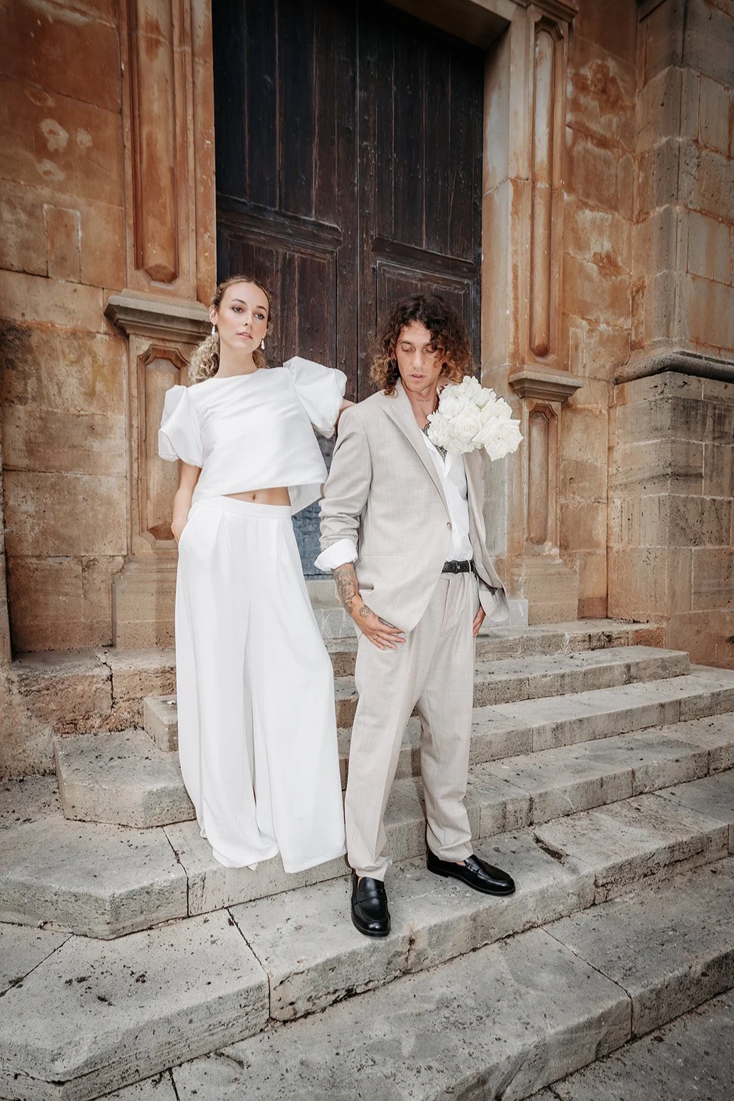 Zwei Menschen stehen auf Steinstufen vor einer alten Holztür, einer hält einen Blumenstrauß aus weißen Blumen, beide tragen elegante, helle Kleidung.