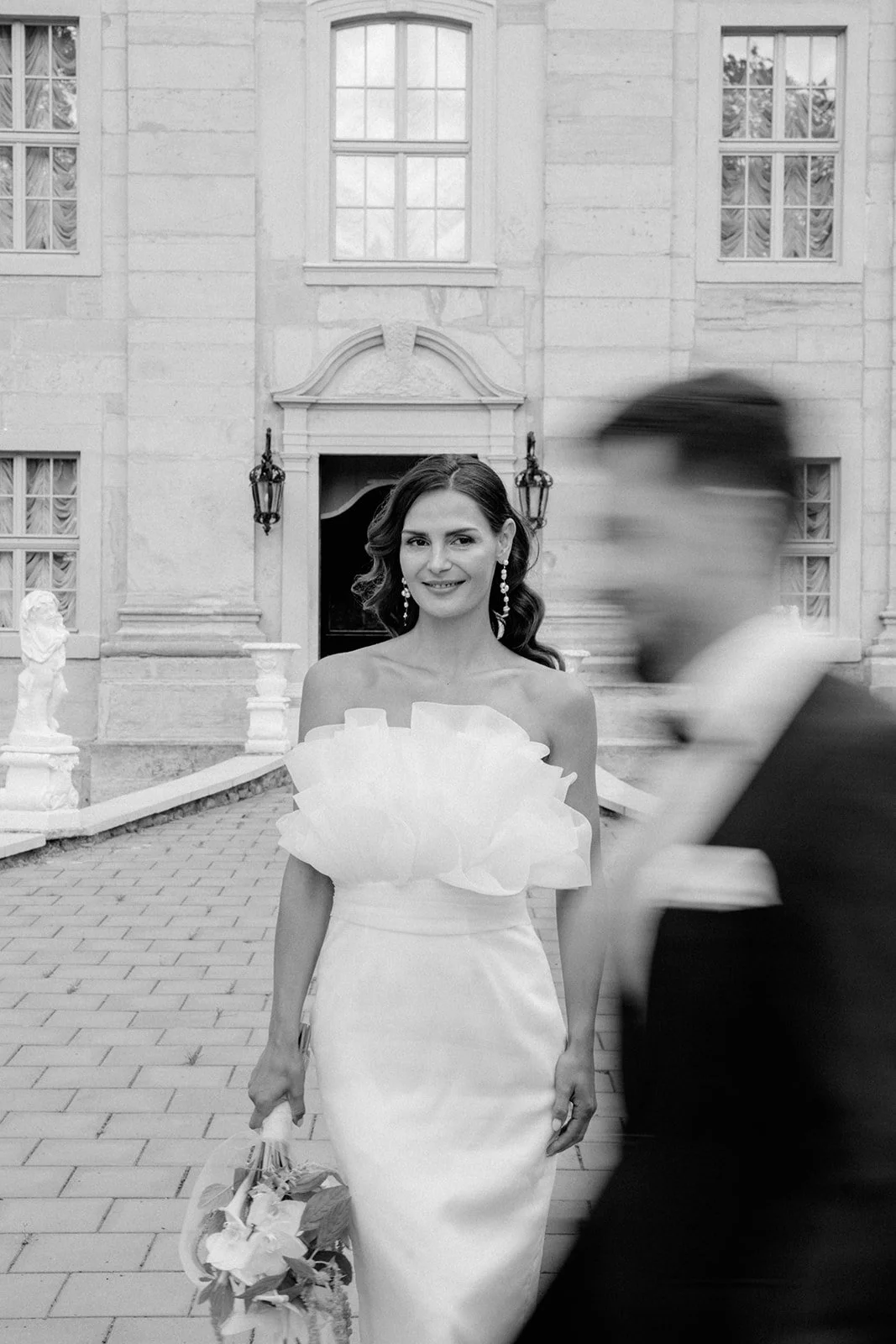 Schwarz-Weiß-Foto einer Hochzeitsbraut in einem eleganten Kleid, die vor einem historischen Gebäude steht, während ein Mann im Anzug im Vordergrund vorbeigeht.