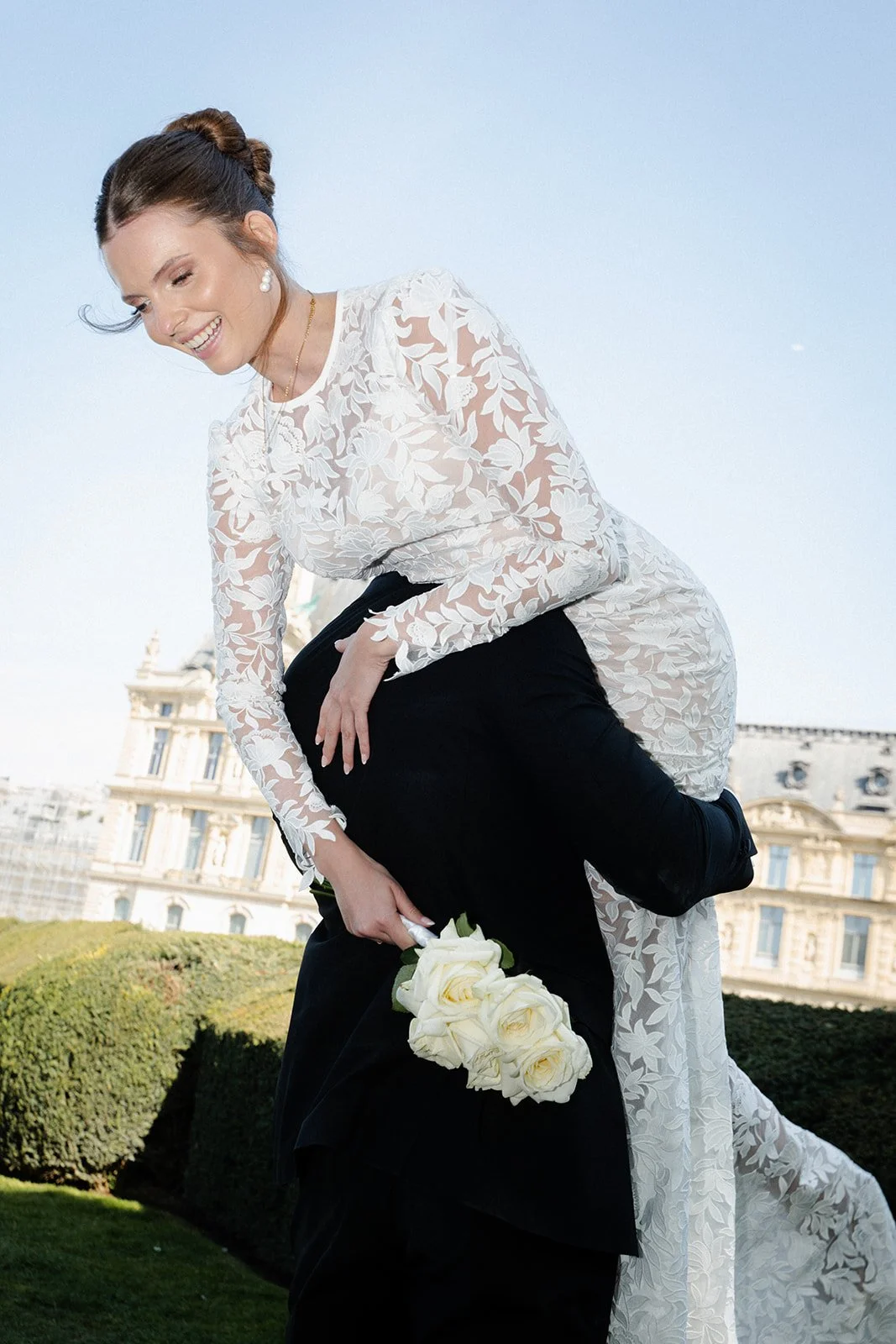 Hochzeitspaar vor einem Gebäude, das im Hintergrund zu sehen ist, die Braut lächelt, hält einen Blumenstrauß, trägt ein weißes Spitzenkleid, der Bräutigam trägt dunklen Anzug.