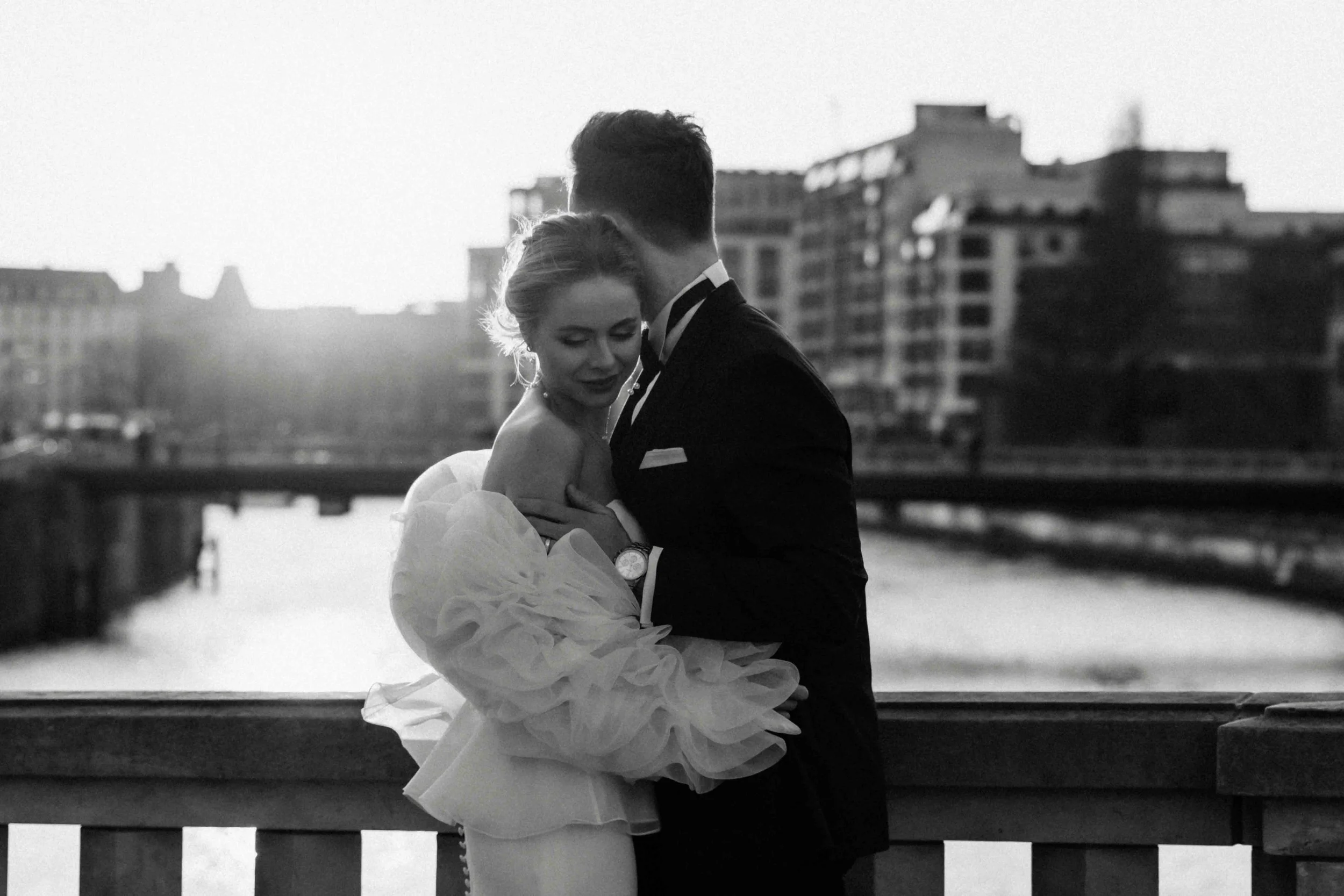 Ein Paar in formeller Kleidung, wahrscheinlich Braut und Bräutigam, sich auf einer Brücke bei Sonnenuntergang in einer Stadt am Fluss, in Schwarz-Weiß-Fotografie.