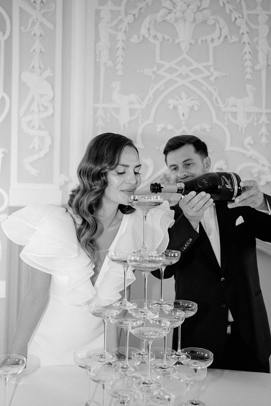 Ein Mann gießt Sekt in ein Cocktailglas, das eine Frau bei einer Feier oder Hochzeit hält, mit einer kunstvoll angeordneten Turm aus leeren Cocktailgläsern.