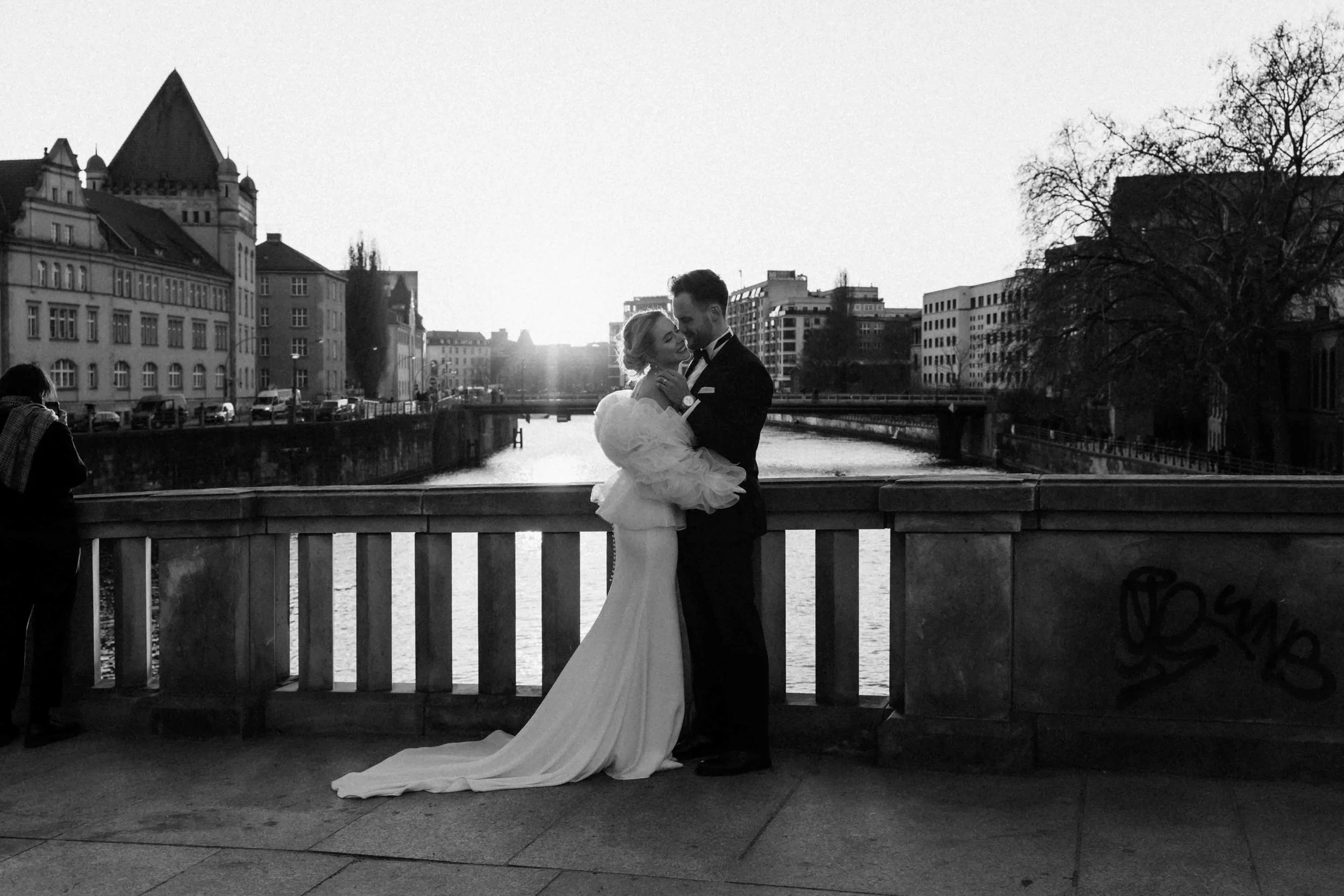 Braut und Bräutigam umarmen sich auf einer Brücke bei Sonnenuntergang in einer Stadt mit Fluss, umgeben von Gebäuden und Bäumen, im schwarz-weißen Stil.