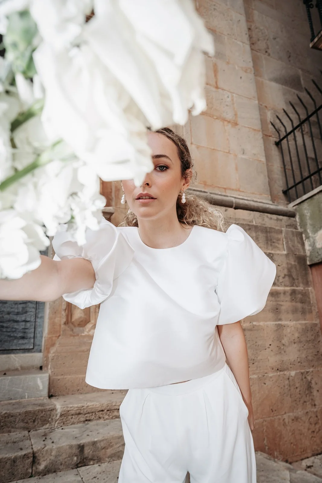 Junge Frau in weißem Kleid posiert vor einer Steintreppe, mit weißen Blumen im Vordergrund.