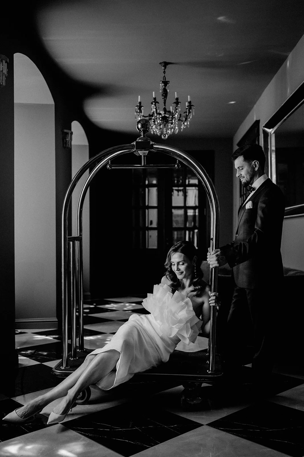 Ein Mann in Smoking schiebt eine Frau in einem Brautkleid auf einem Hotelwagen durch ein elegantes Hotel. Das Bild ist schwarz-weiß und zeigt einen Kristallleuchter an der Decke.