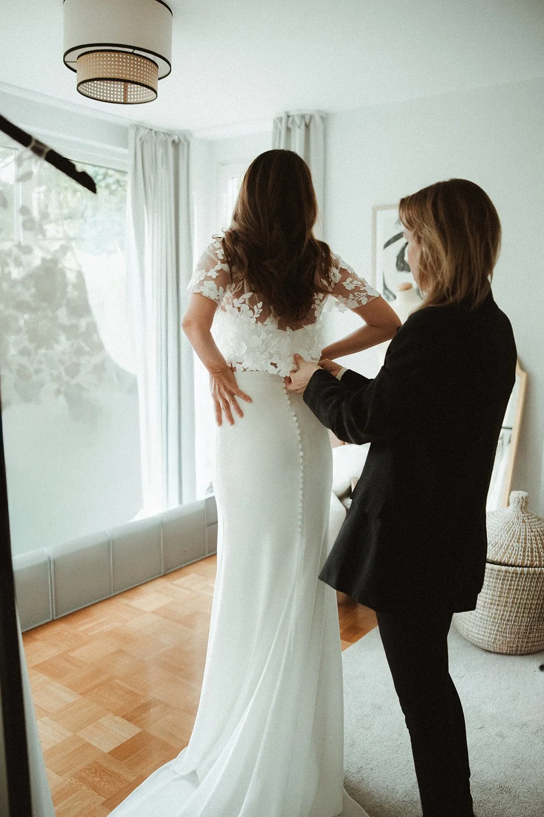 Braut wird angezogen. Sie trägt ein langes weißes Brautkleid mit Spitzenärmeln. Eine andere Person hilft ihr beim Anziehen.