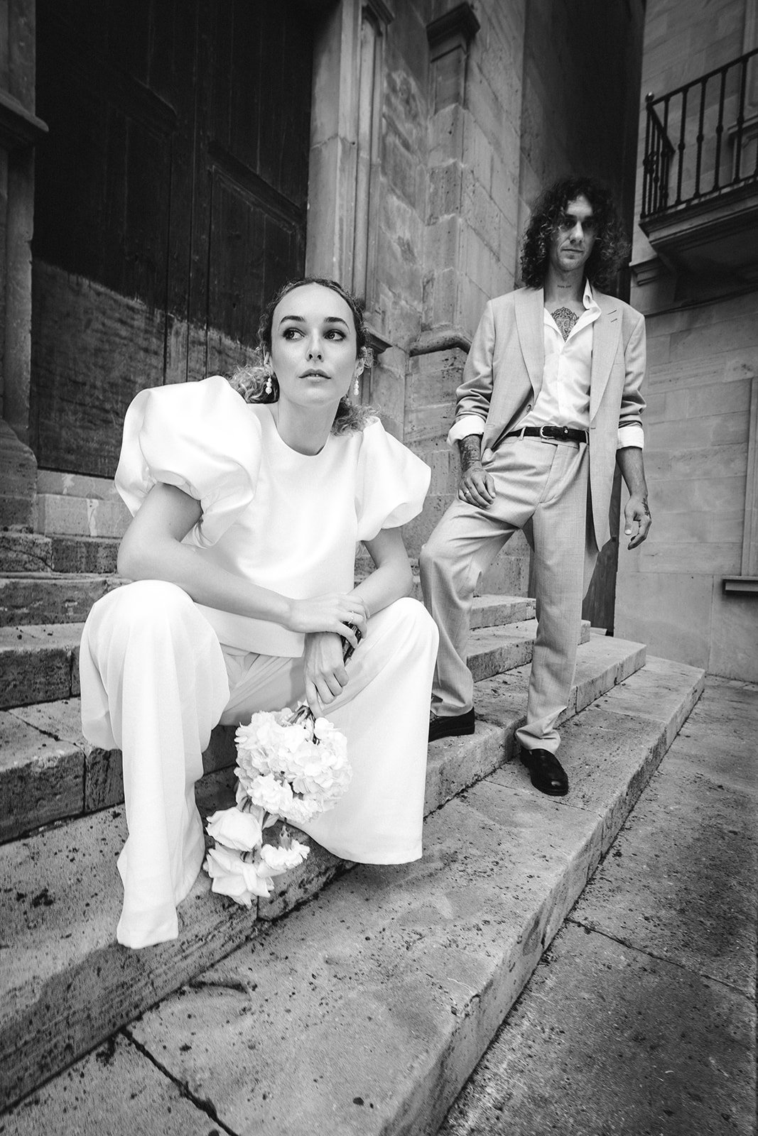 Schwarz-weiß-Foto von einer Frau in weißem Kleid mit Puffärmeln, die auf den Steinen einer Treppe sitzt, und einem Mann in einem hellen Anzug, der an der Seite der Treppe steht. Sie halten Blumen in der Hand.