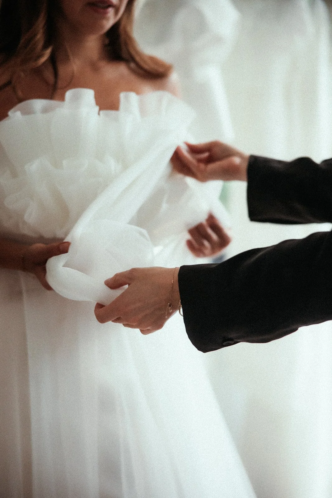 Nahaufnahme einer Person im Hochzeitskleid, während ein anderer Mensch den Stoff des Kleides arrangiert oder hält. Der Fokus liegt auf den Händen und dem Stoff.