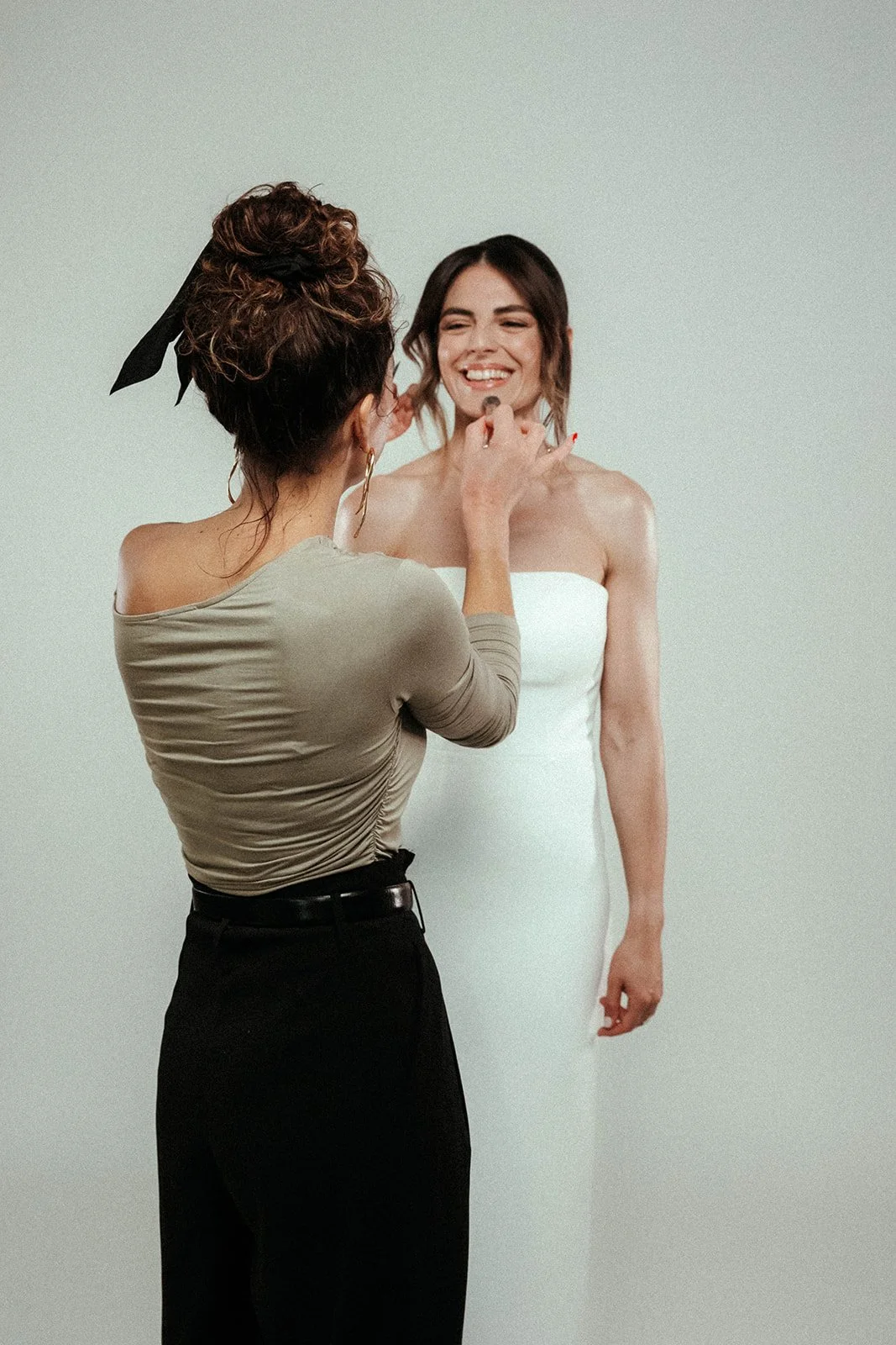 Eine Frau wird für ein Fotoshooting mit Make-up vorbereitet, trägt ein weißes Kleid, während eine Visagistin sie schminkt, vor einem neutralen Hintergrund.
