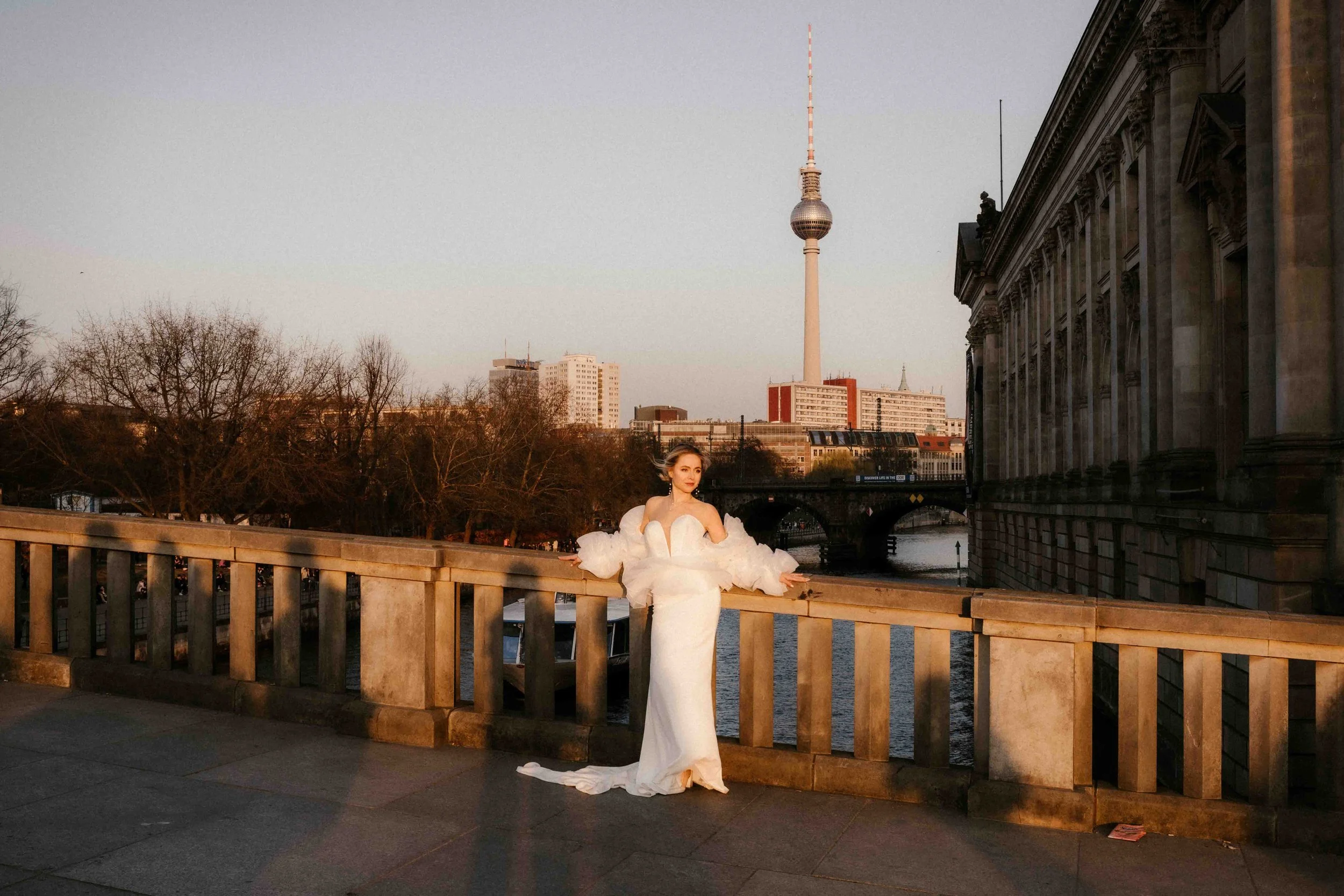 Eine Frau in einem eleganten weißen Hochzeitskleid steht an einem Flussufer mit Berliner Fernsehturm im Hintergrund, bei Sonnenuntergang.