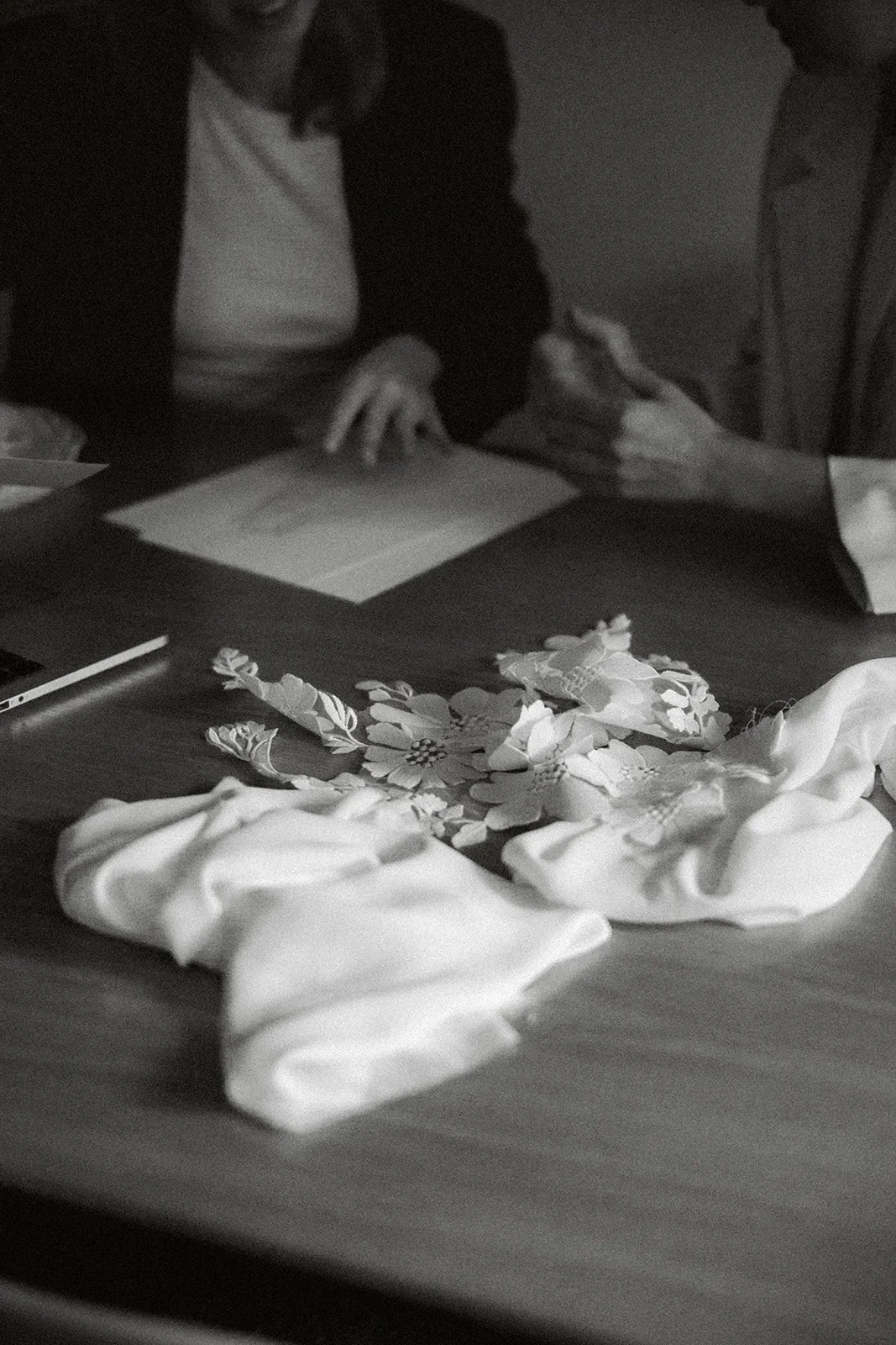 Schwarzweißfoto von zwei Personen, die an einem Tisch sitzen, mit Stoffmustern und Blumenapplikationen auf der Tischoberfläche.