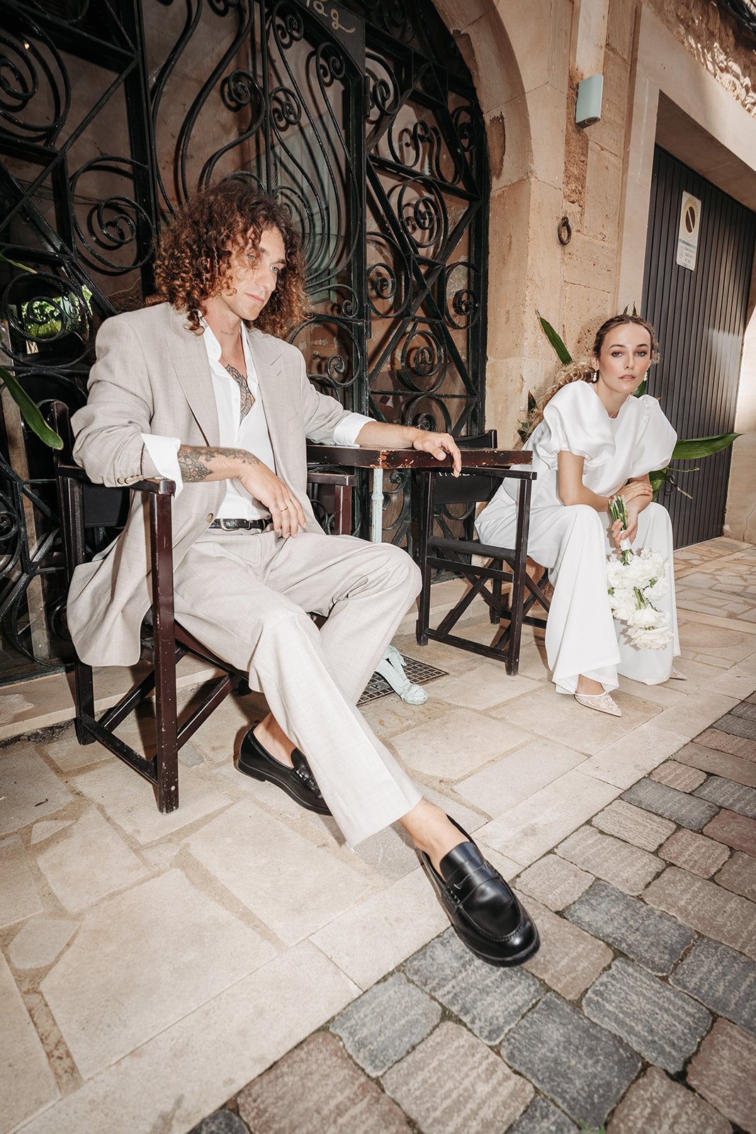 Ein Mann und eine Frau sitzen auf einer Bank und einem Stuhl vor einer Steinzäune, die an einem alten Gebäude befestigt sind. Der Mann trägt einen beigen Anzug und schwarze Schuhe, während die Frau in einem weißen Kleid sitzt und einen Blumenstrauß i
