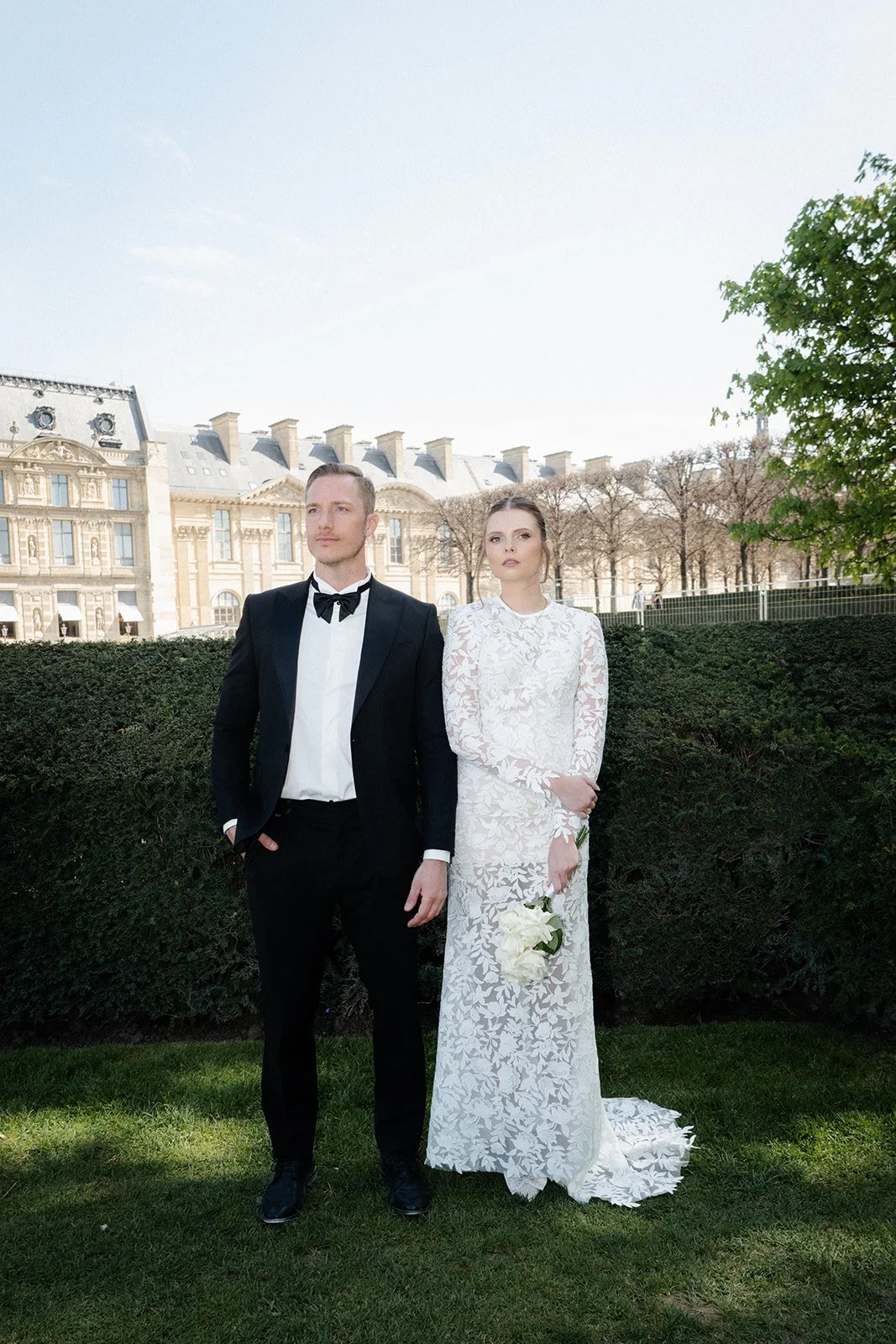 Ein Mann in einem schwarzen Anzug mit Fliege und eine Frau in einem weißen Spitzenkleid, die weiße Blumen hält, stehen auf einem Rasen vor einem Büsche, im Hintergrund ein historisches Gebäude.