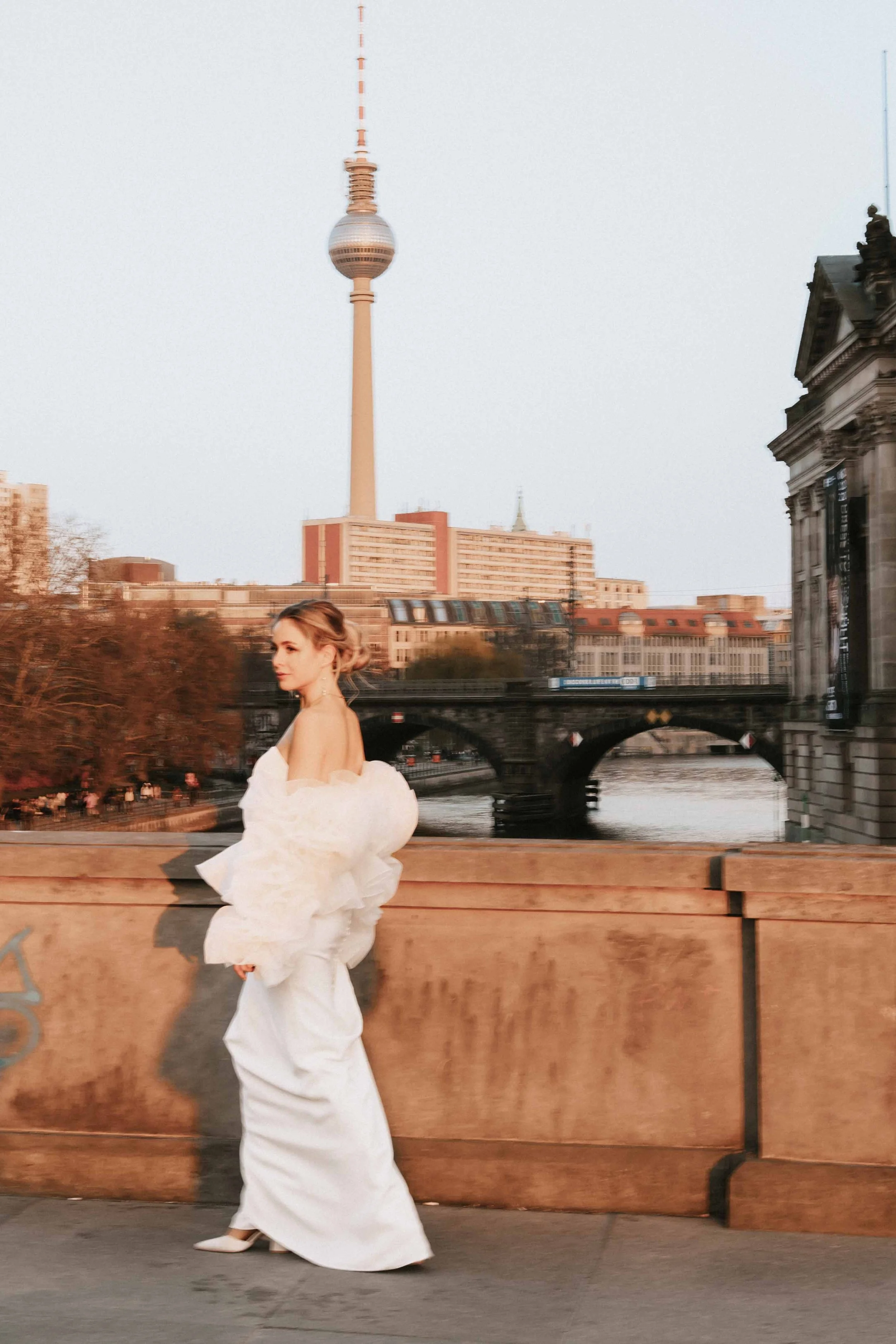 Frau in weißem Kleid auf einer Brücke in Berlin, mit Fernsehturm im Hintergrund bei Sonnenuntergang.