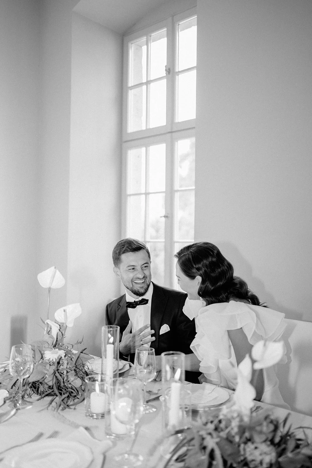 Ein Lächelndes Paar bei einem eleganten Essen in einem hellen Raum mit großen Fenstern, festlich gedeckter Tisch mit Kerzen und Blumen, Schwarz-Weiß-Fotografie.