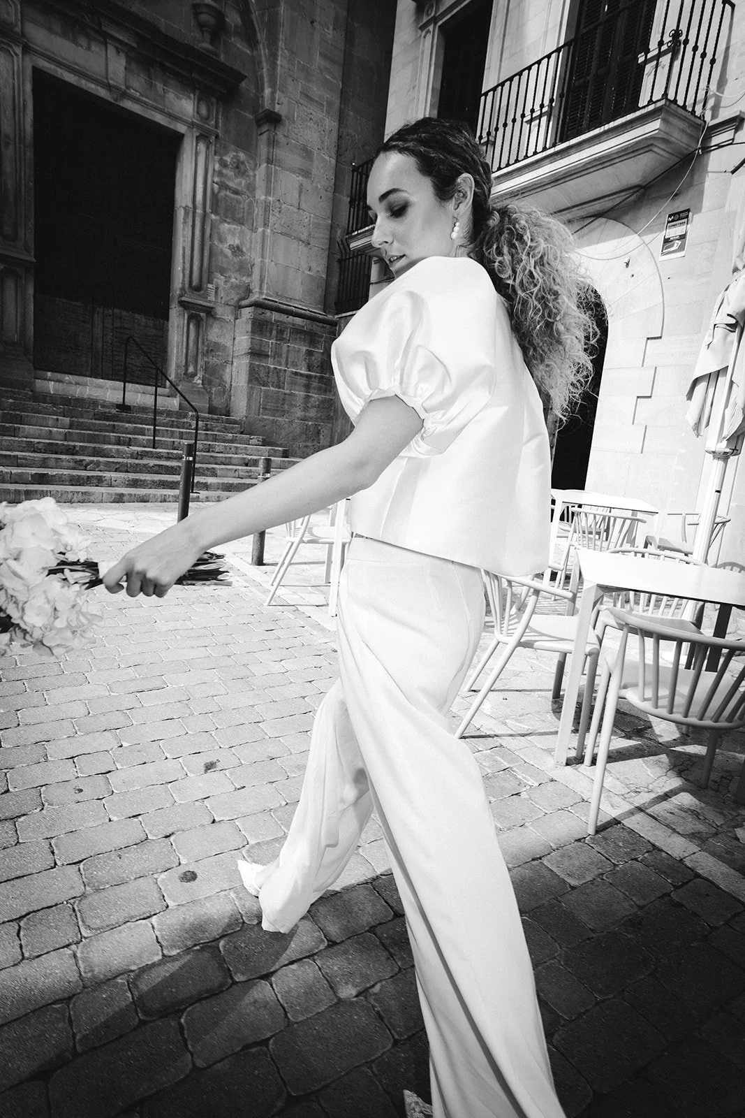 Frau mit dunklen, lockigen Haaren und hellen Ohrringen, trägt eine weiße, puffärmelige Bluse und weiße weite Hosen, hält einen Blumenstrauß, steht auf einer Kopfsteinpflasterstraße im Freien.