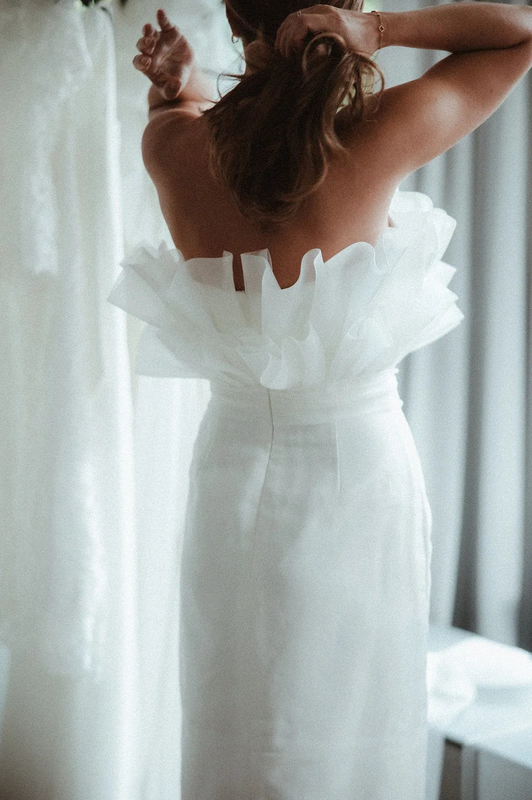 Rückansicht einer Frau in einem weißen Hochzeitskleid mit Rüschen, die sich die Haare hochbindet.