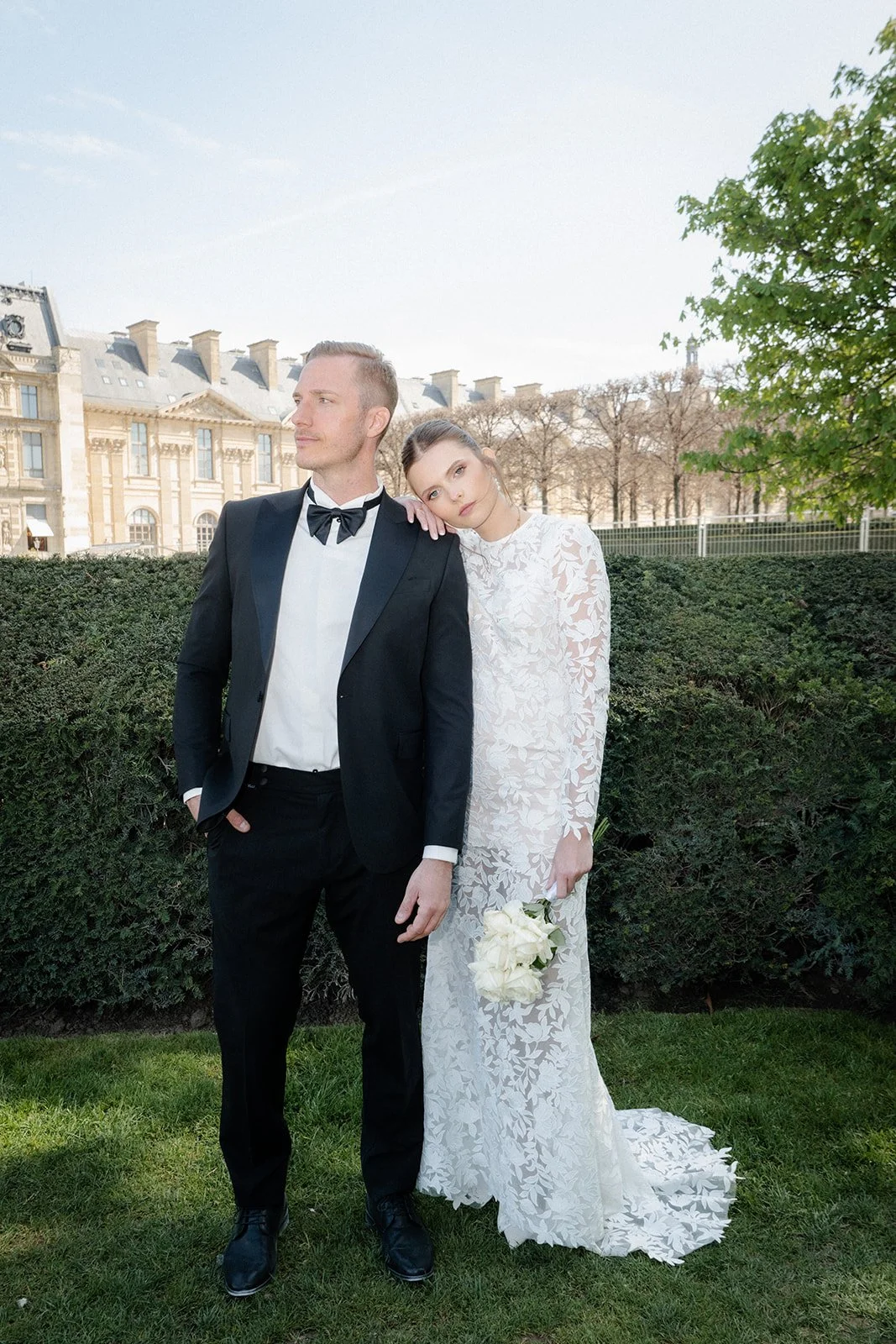Ein frisch verheiratetes Paar steht im Grünen, das Brautkleid ist weiß mit floralem Muster, sie hält einen Brautstrauß, er trägt einen schwarzen Anzug mit Fliege.