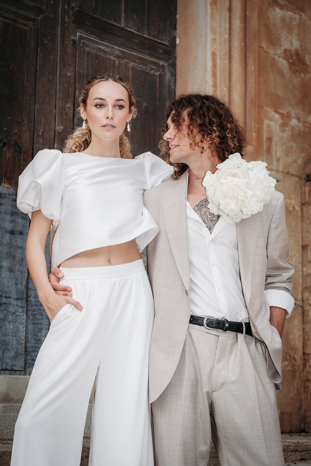 Ein Paar in formeller Kleidung steht vor einer alten Steinmauer. Die Frau trägt einen weißen, kurzen, puffy Oberteil und lange Hosen mit offenen Taschen, während der Mann einen hellgrauen Anzug mit einem weißen Hemd und einer schwarzen Gürtel trägt. 