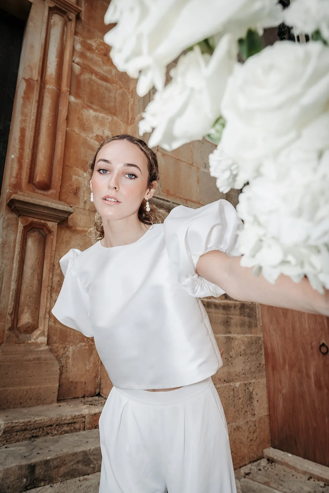 Junge Frau in weißem Oberteil und Hose steht vor einer Ziegelwand, macht ein Selfie und hält eine große weiße Blumenarrangement.