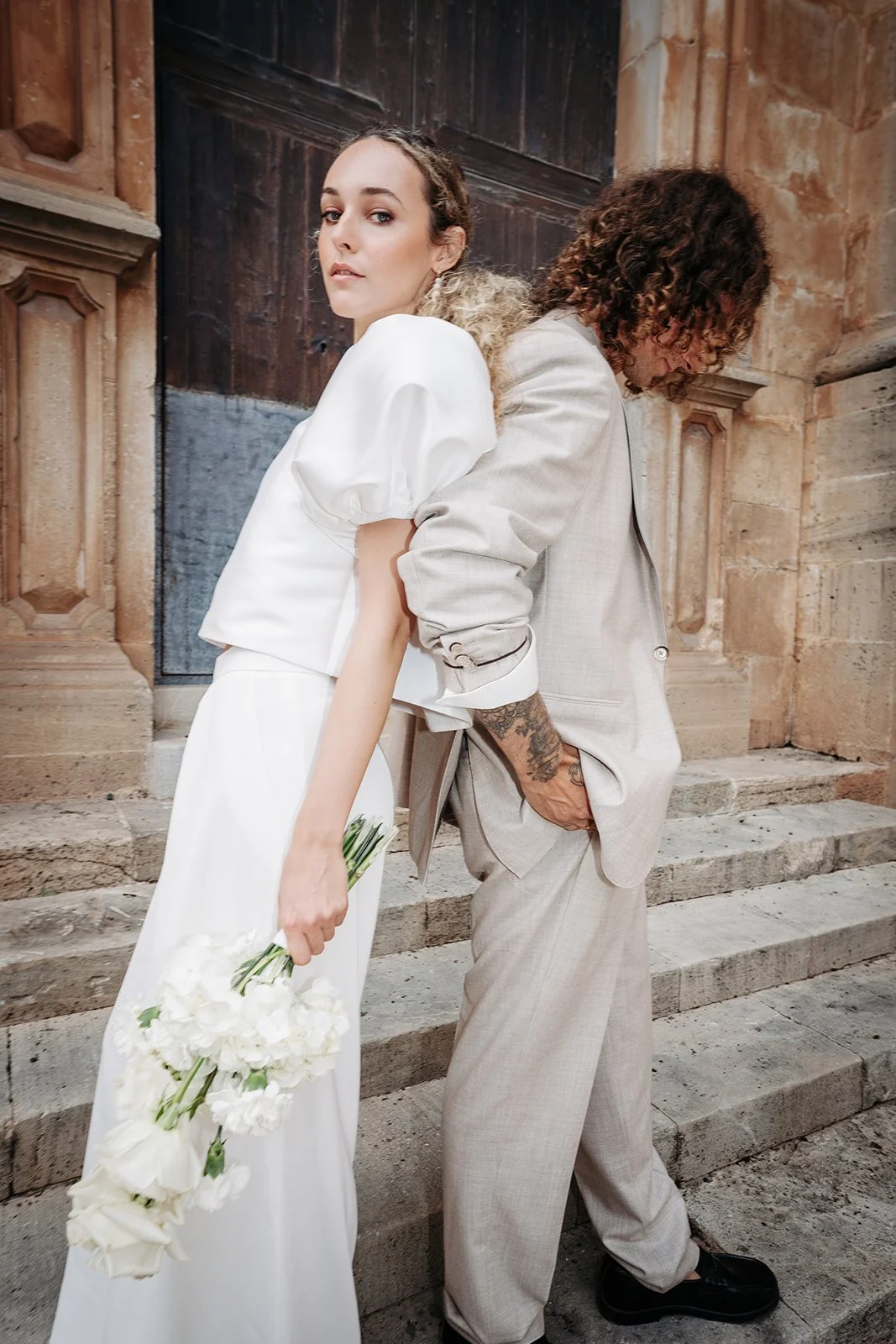 Ein Mann und eine Frau stehen Rücken an Rücken vor einer alten Steinmauer und Treppe. Die Frau trägt ein weißes Kleid und hält einen Blumenstrauß. Der Mann trägt einen hellgrauen Anzug und hat Tattoos am Arm.