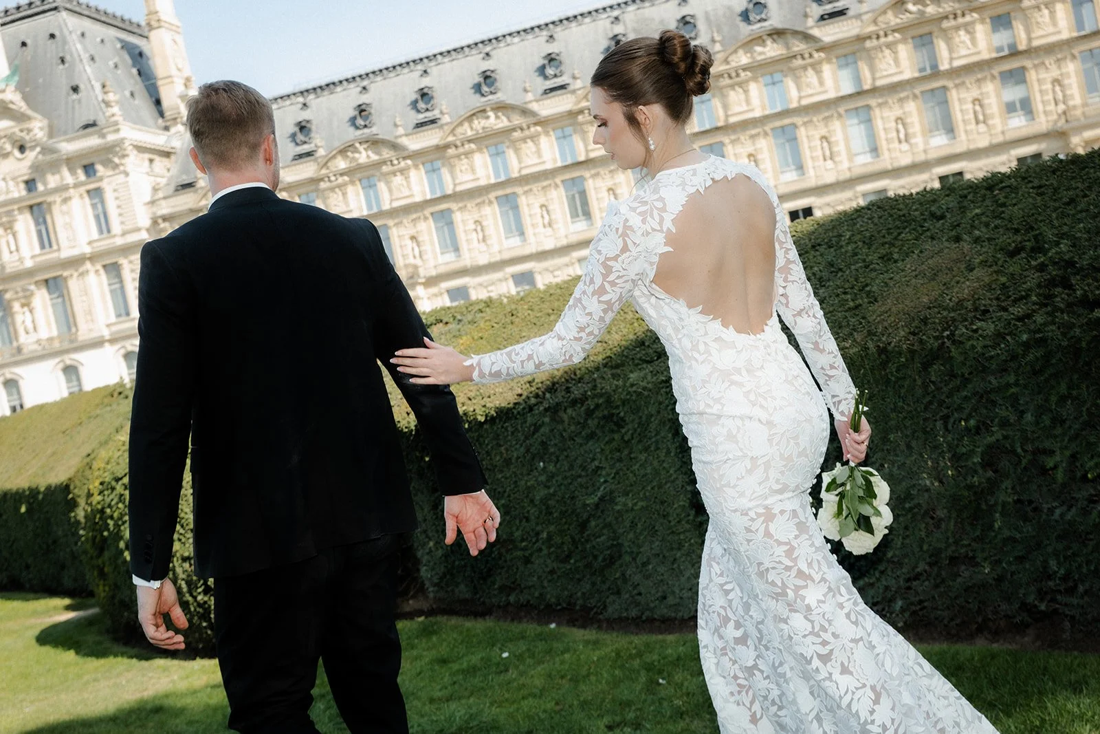 Ein Brautpaar, eine Frau in einem weißen Hochzeitskleid und ein Mann in einem schwarzen Anzug, schlendert auf einer Rasenfläche vor einem historischen Gebäude mit Jugendstilarchitektur, während die Braut den Mann berührt.