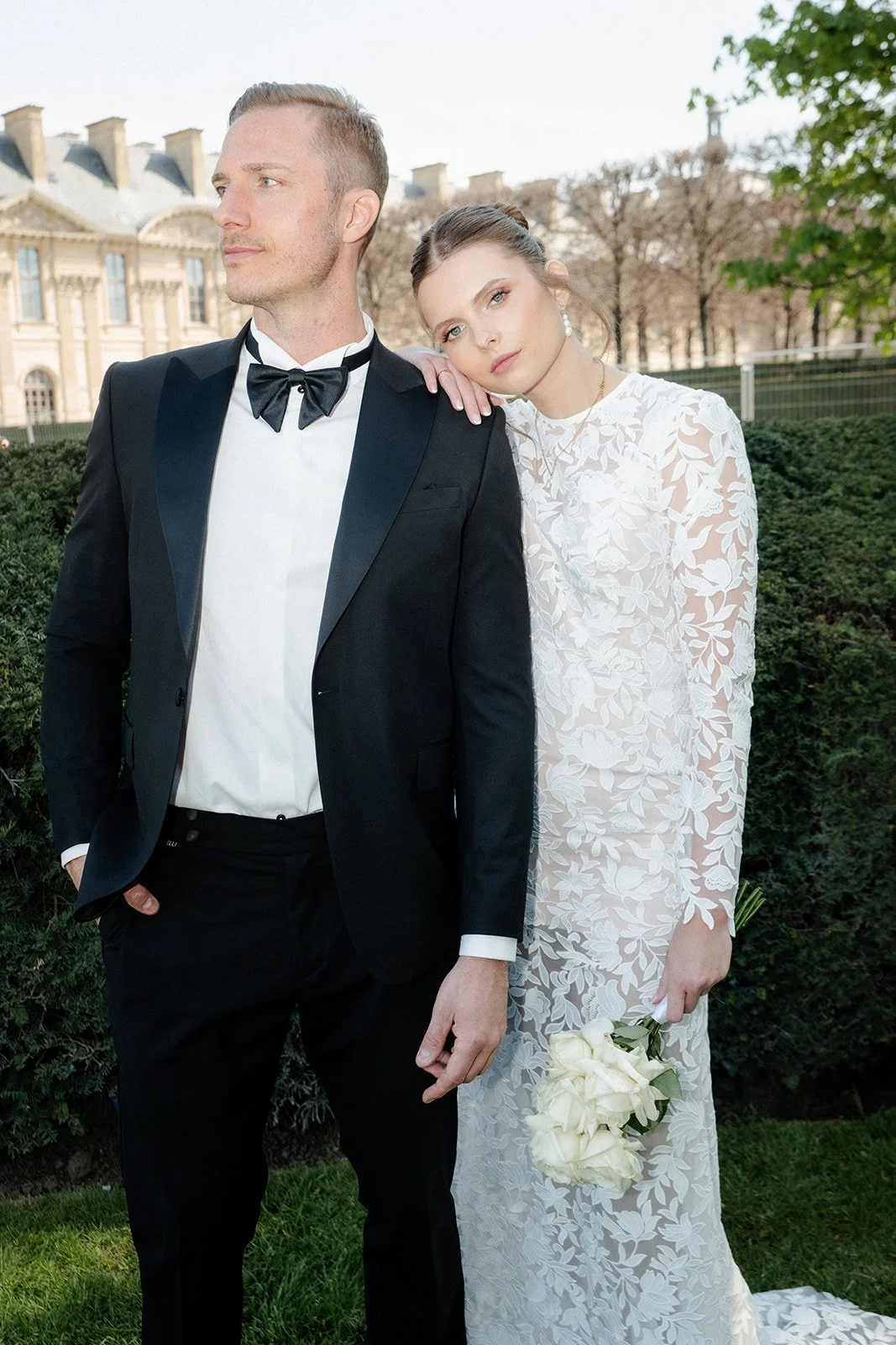 Ein Paar in formeller Kleidung vor einem Garten, das Mädchen in einem weißen Brautkleid mit Blumenmuster hält einen Blumenstrauß, der Junge trägt einen schwarzen Anzug mit Fliege.