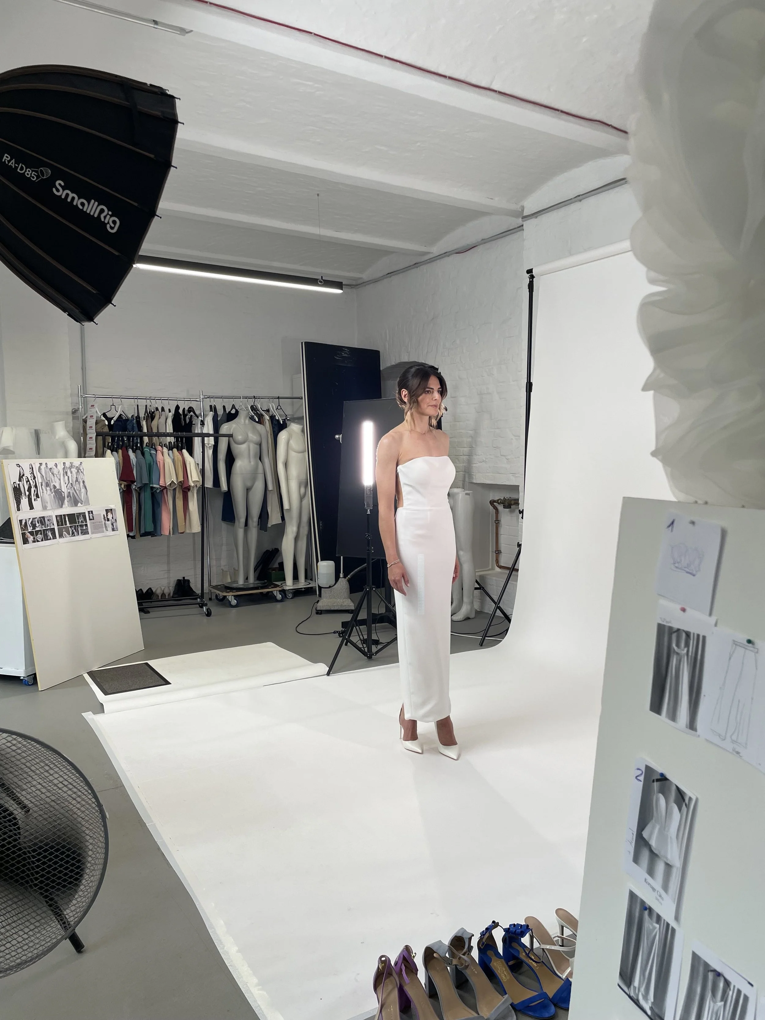 Eine Frau posiert in einem weißen Brautkleid vor einem weißen Fotohintergrund in einem Fotostudio mit Fotografieausrüstung und Kleidungsstücken im Hintergrund.