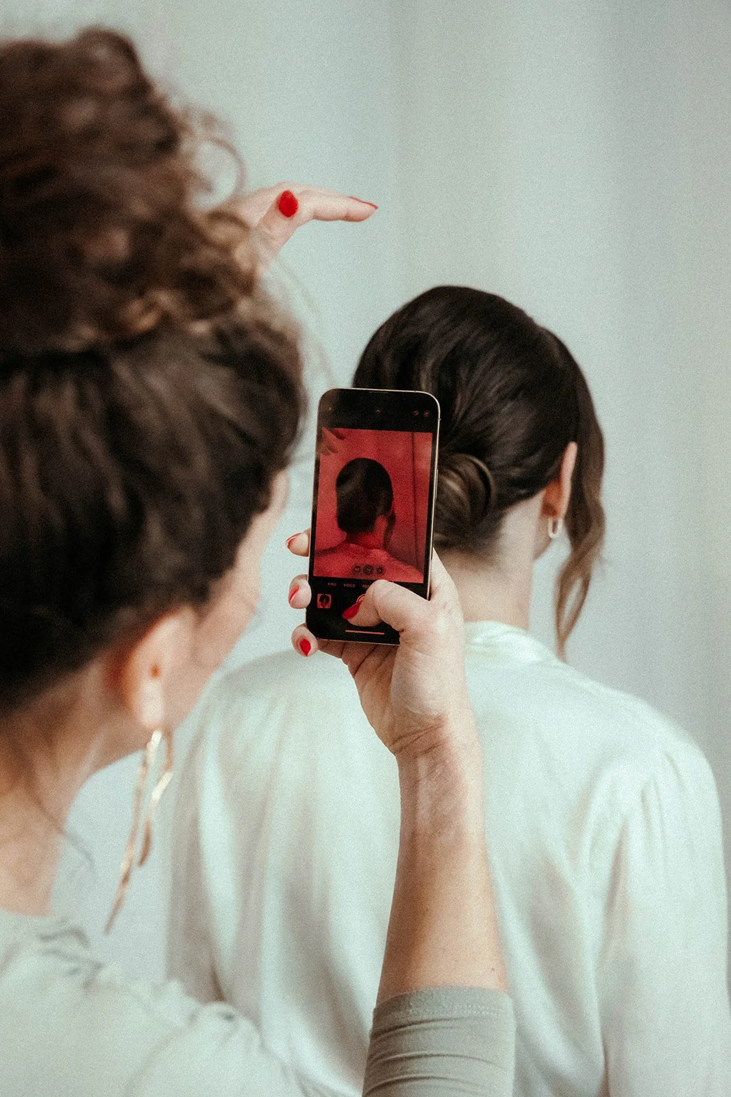 Eine Frau macht ein Foto der Frisur einer anderen Frau mit einem Smartphone. Beide Frauen haben dunkle Haare und tragen helle Kleidung.