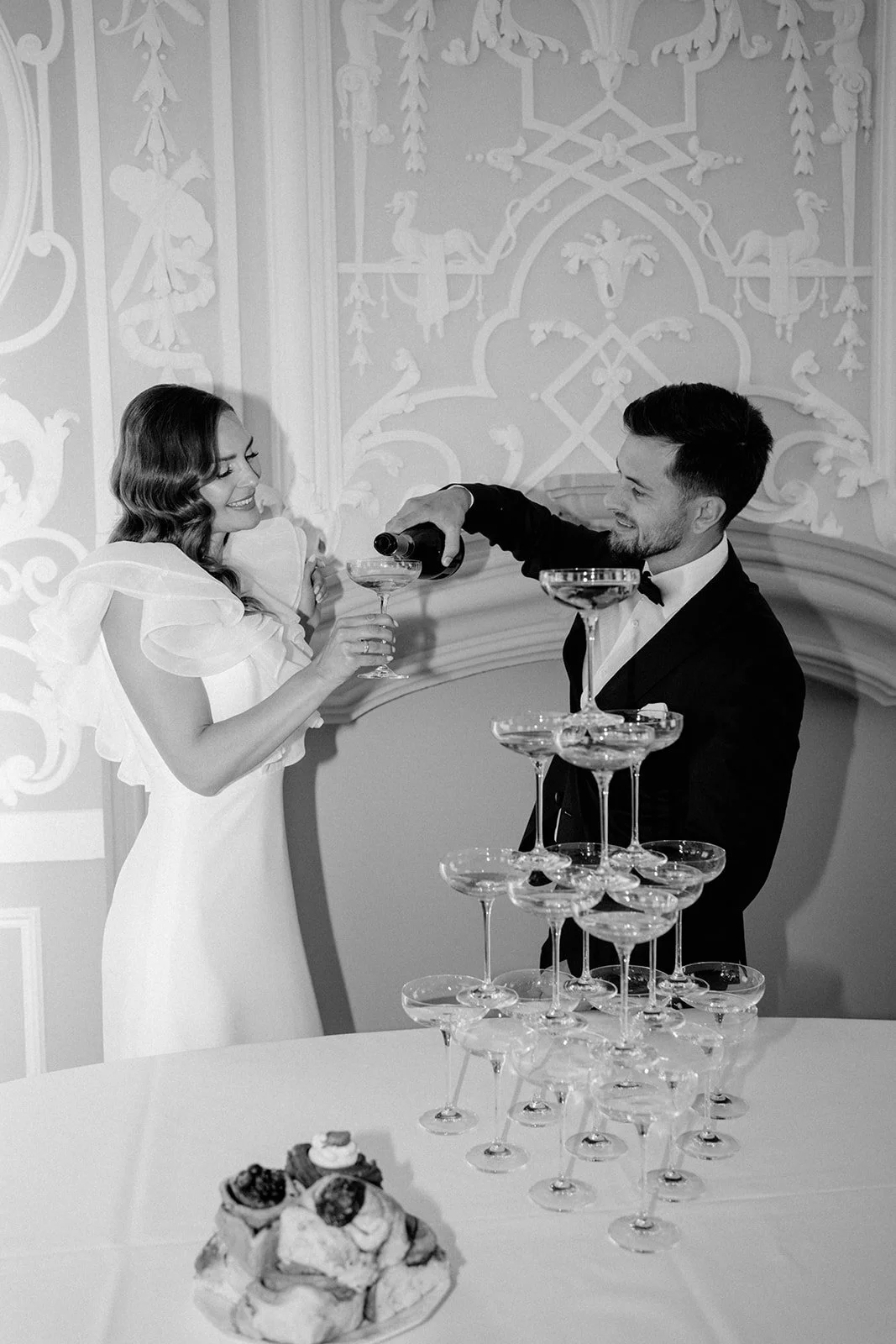 Ein Paar bei einem Hochzeitsempfang, der Bräutigam schenkt der Braut einen Champagner. Es gibt ein Turm mit leeren Champagnergläsern auf einem Tisch und eine Süßigkeitenplatte im Vordergrund.