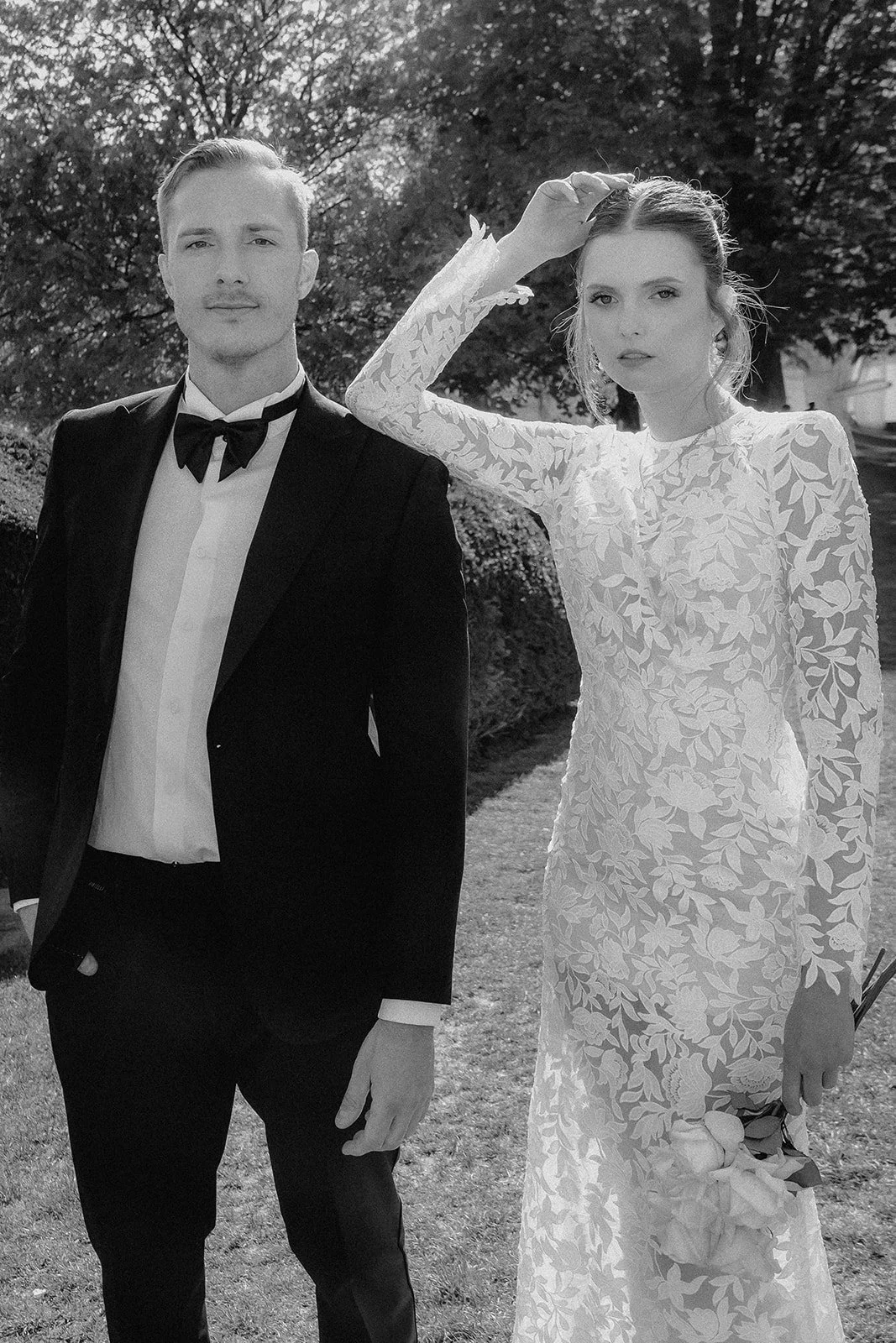 Schwarz-weiß Foto eines Paares in formeller Kleidung im Freien, Frau trägt ein langärmeliges, floral gemustertes Kleid, Mann trägt einen Smoking mit Fliege.