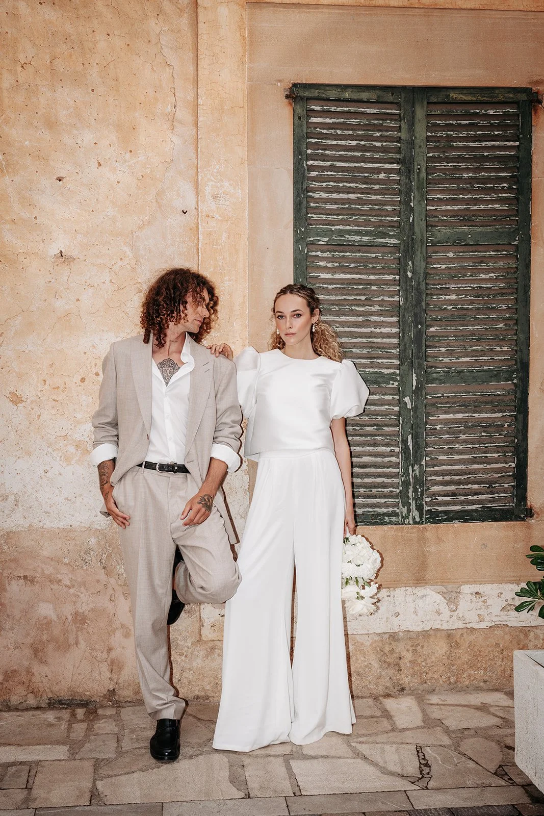 Zwei Personen, eine Frau in weißen Kleid mit Blumen und einen Mann in Anzug, stehen vor einer alten Hauswand mit geschlossenem Fensterläden.