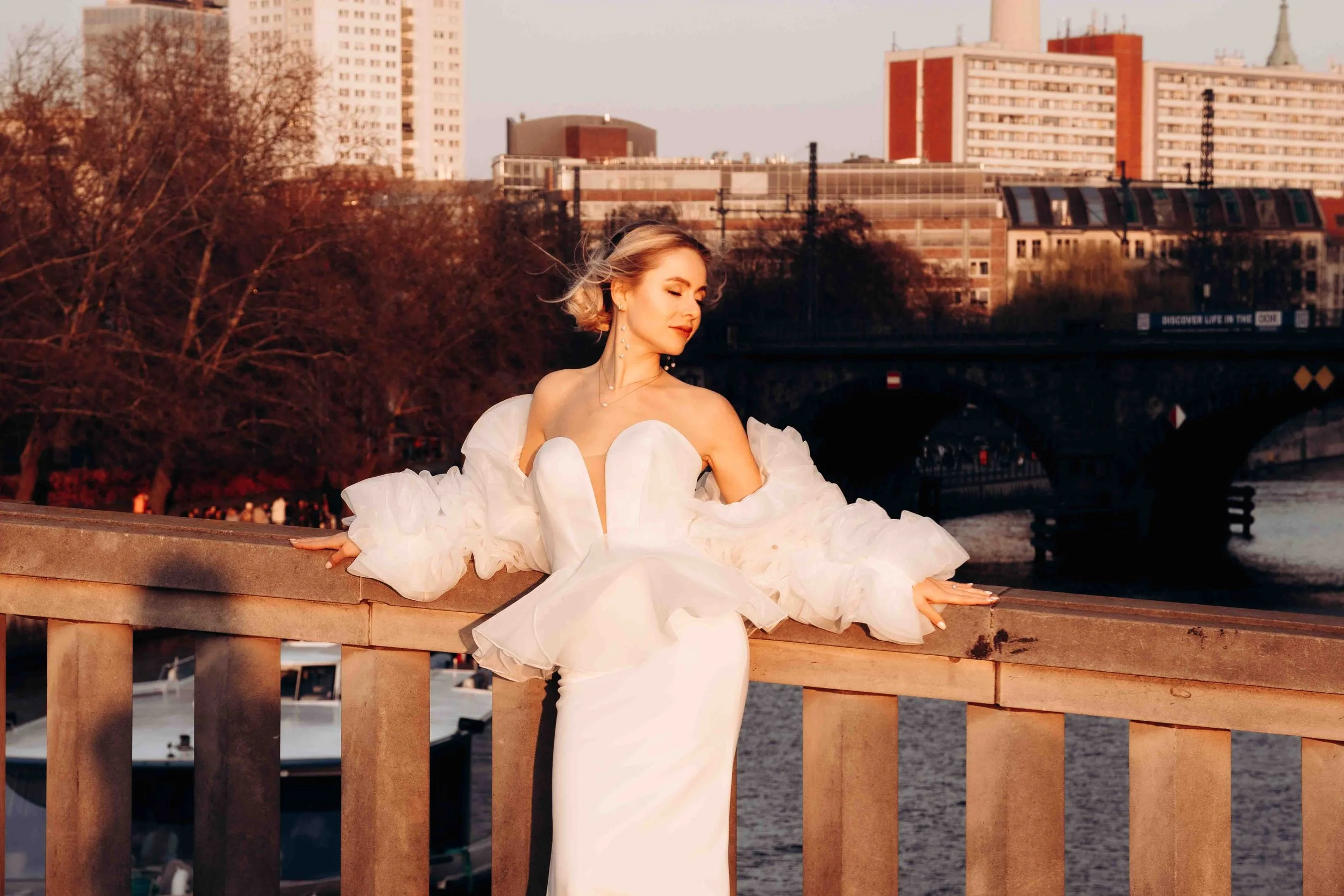 Frau in einem eleganten weißen Abendkleid auf einer Brücke am Fluss bei Sonnenuntergang, mit Stadtgebäuden im Hintergrund.