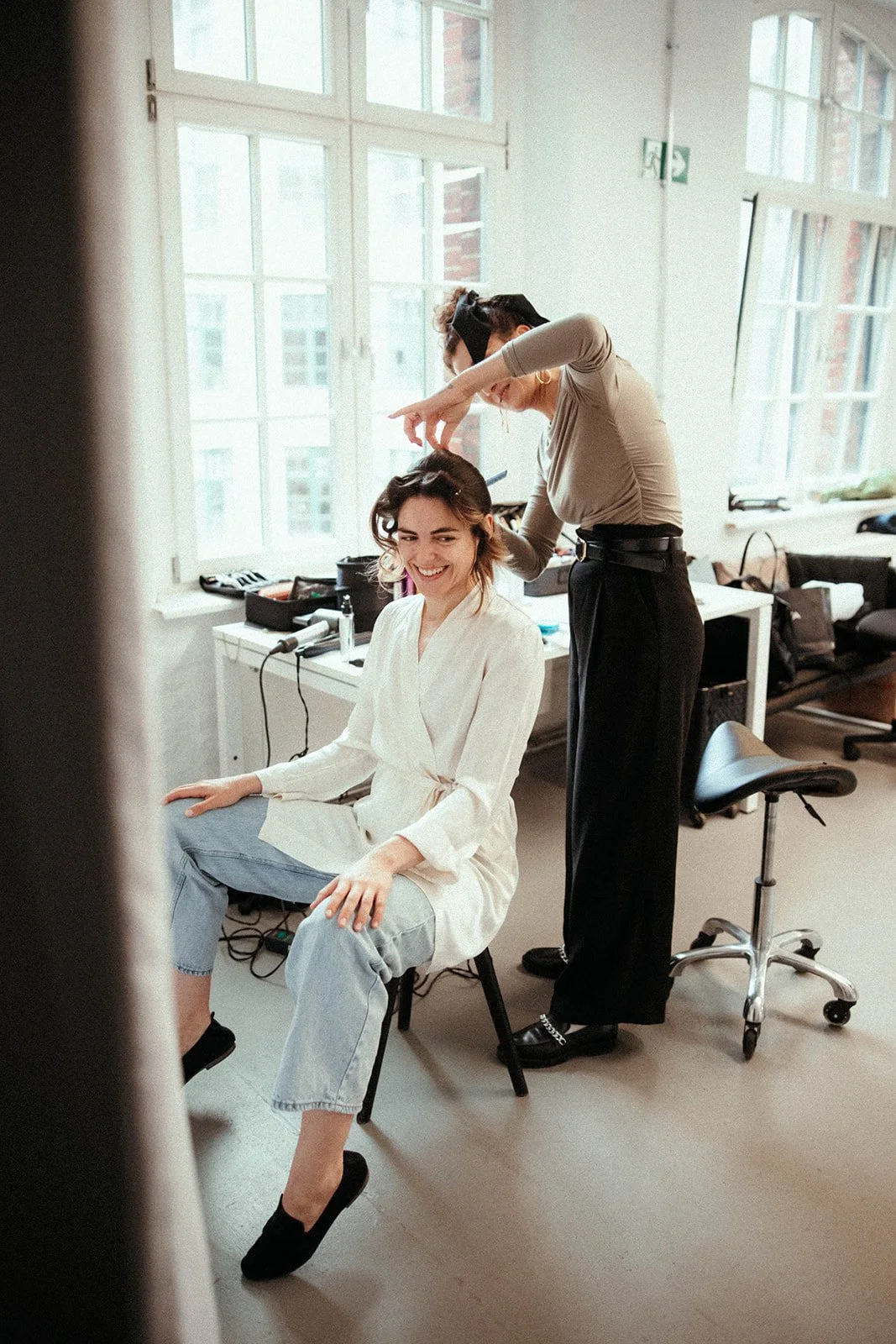 Eine Frau sitzt lachend auf einem Stuhl, während eine Friseurin sie frisiert. Das Bild ist in einem schön beleuchteten Raum mit großen Fenstern aufgenommen.